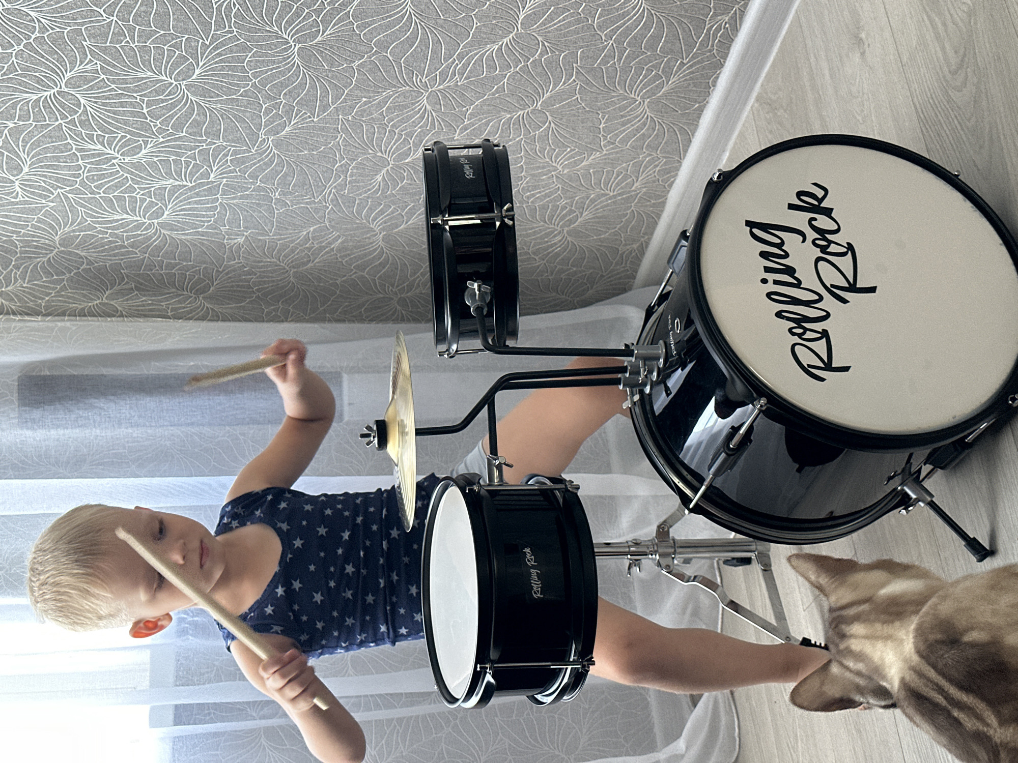 A young Caucasian boy with blond hair is playing a small drum set. He is wearing a navy blue tank top with white stars. He is holding drumsticks in both hands and appears to be striking the drums. The drum set is black and has the brand name "Rolling Rock" printed on it. A cat with tabby markings is looking at the drum set. The scene appears to be indoors, possibly in a residential setting, with white sheer curtains in the background and patterned wallpaper. The lighting suggests it is daytime. This image does not provide enough information to determine the location in the city of Energetik, Russia.
