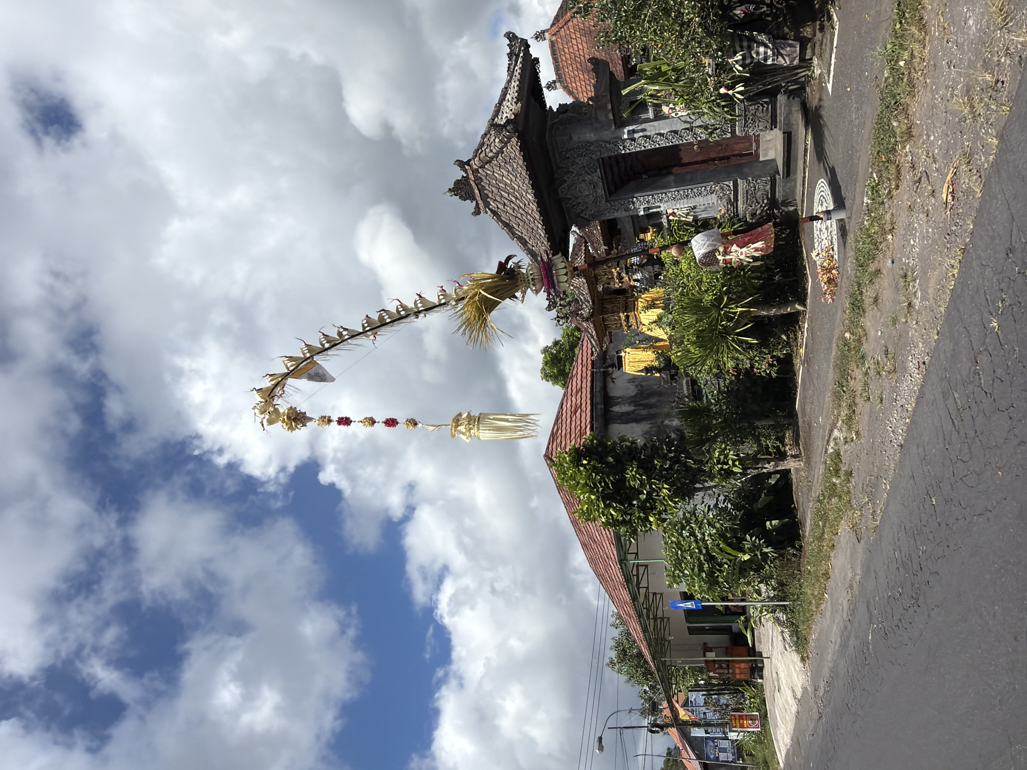 In Kintamani, Indonesia, a street scene unfolds under a partly cloudy sky. A long, decorative pole, adorned with golden ornaments and red flowers, arches gracefully over the frame. In the foreground, a paved road shows signs of wear, with patches of dry grass and gravel lining its edge. To the right, a traditional Balinese gate, intricately carved and topped with terracotta roof tiles, stands guard. A person in a red and white garment is visible near the gate, holding a similar decorative item. Beyond the gate, lush green foliage and a building with a red-tiled roof are visible. To the left, another building with a red-tiled roof and a covered walkway is situated along the street, with a blue pedestrian crossing sign nearby. The overall impression is of a vibrant cultural setting in a bustling Indonesian town.
