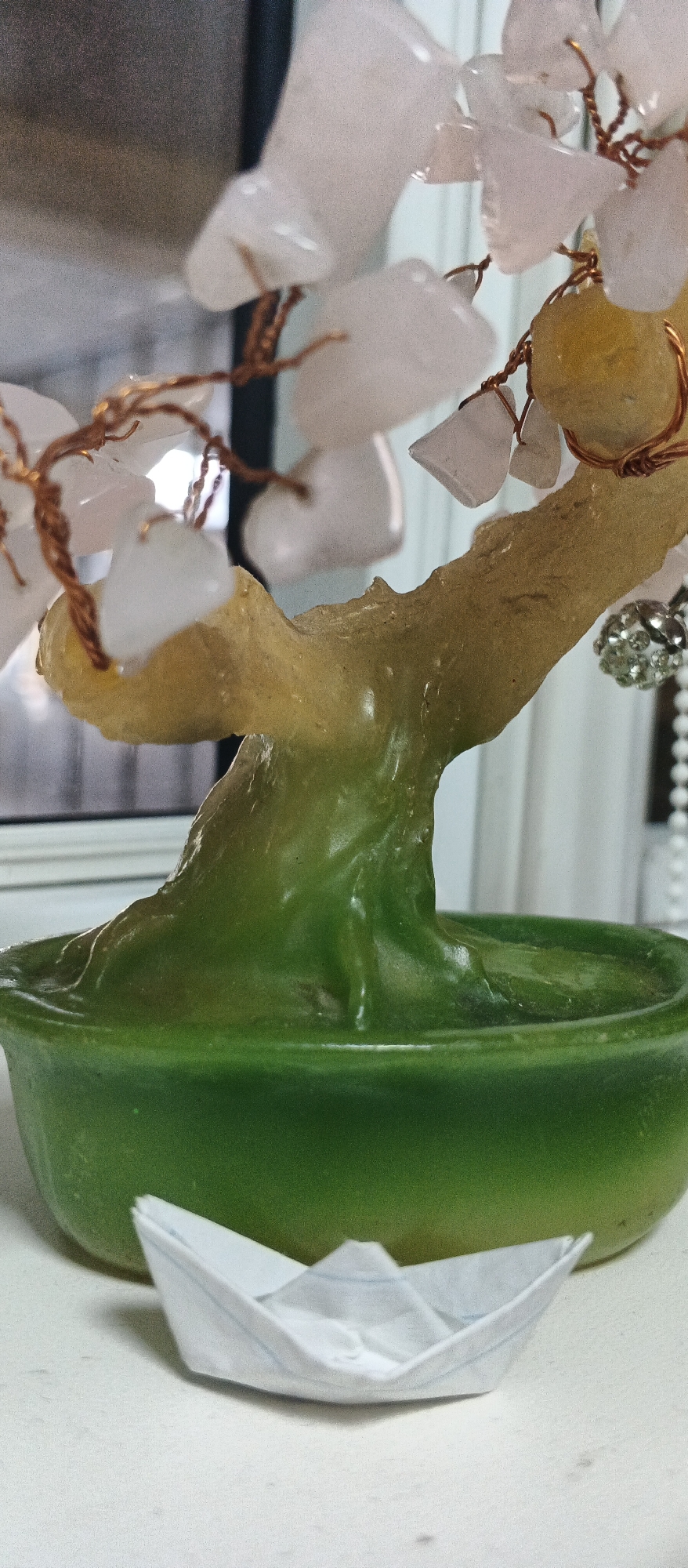 A close-up, vertical, eye-level shot shows a decorative tree made of light pink and yellow stones with copper wire branches. The tree is in a green, round bowl that contains a green, wavy, resinous base. In the foreground, a small, white paper boat is placed on a white surface. The background is out of focus, showing a dark surface on the left and vertical light-colored lines on the right, possibly indicating a window or blinds. There are no people visible in the image, and no specific location in Smila, Ukraine can be determined from the visual information.