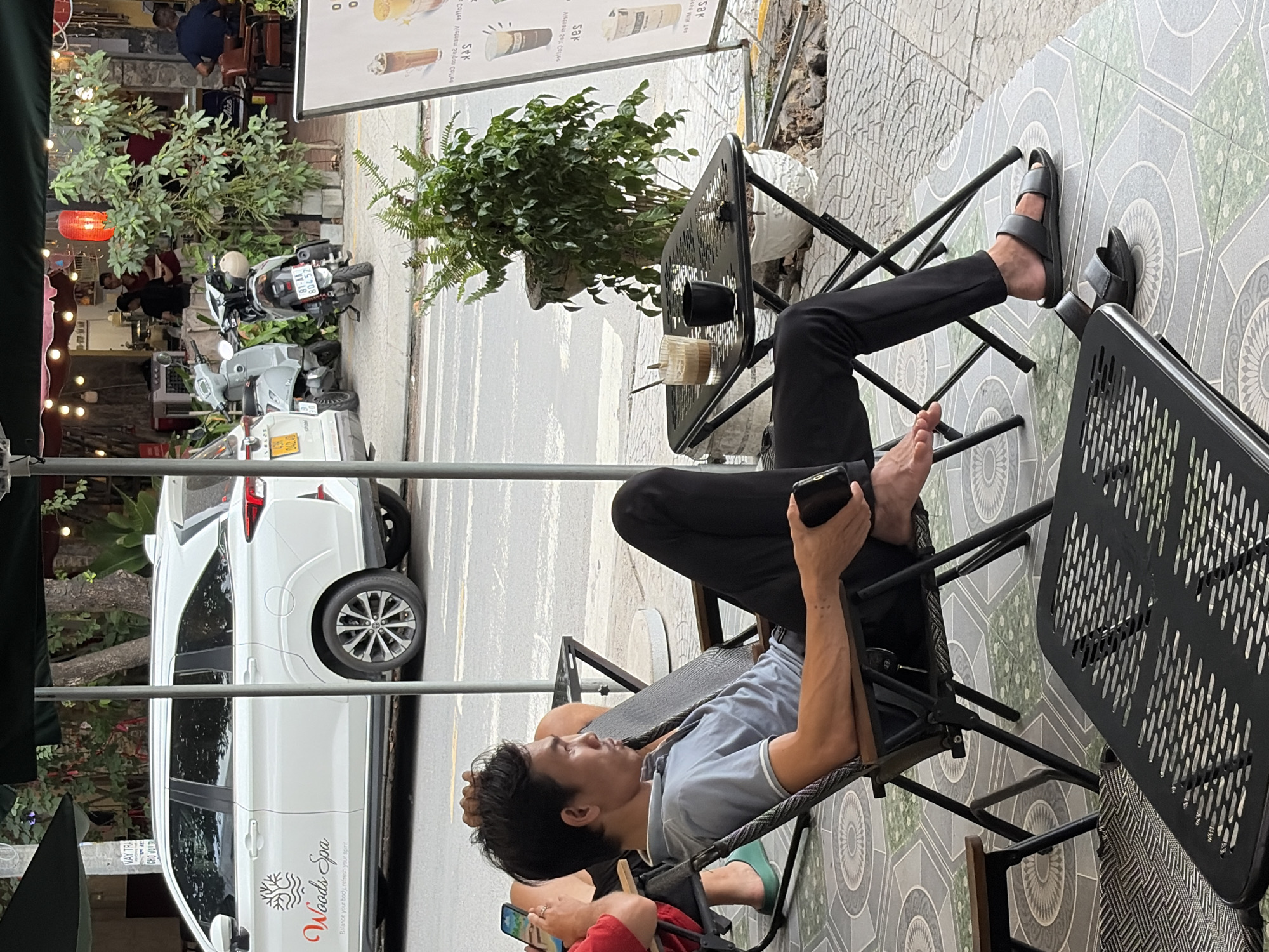 A man sits in a black folding chair at a table outside a cafe in Đà Nẵng, Vietnam. He wears a light blue collared shirt and black pants. The man is holding a smartphone in his right hand and has his left hand resting on his head. In front of him on the table are a glass of iced coffee and a small black cup. The cafe has a tiled floor and is decorated with potted plants. In the background, a white SUV with the logo "Woods Spa" is parked on the street, and several motorcycles are visible. The scene is illuminated by daylight, with some overhead lighting from the cafe.