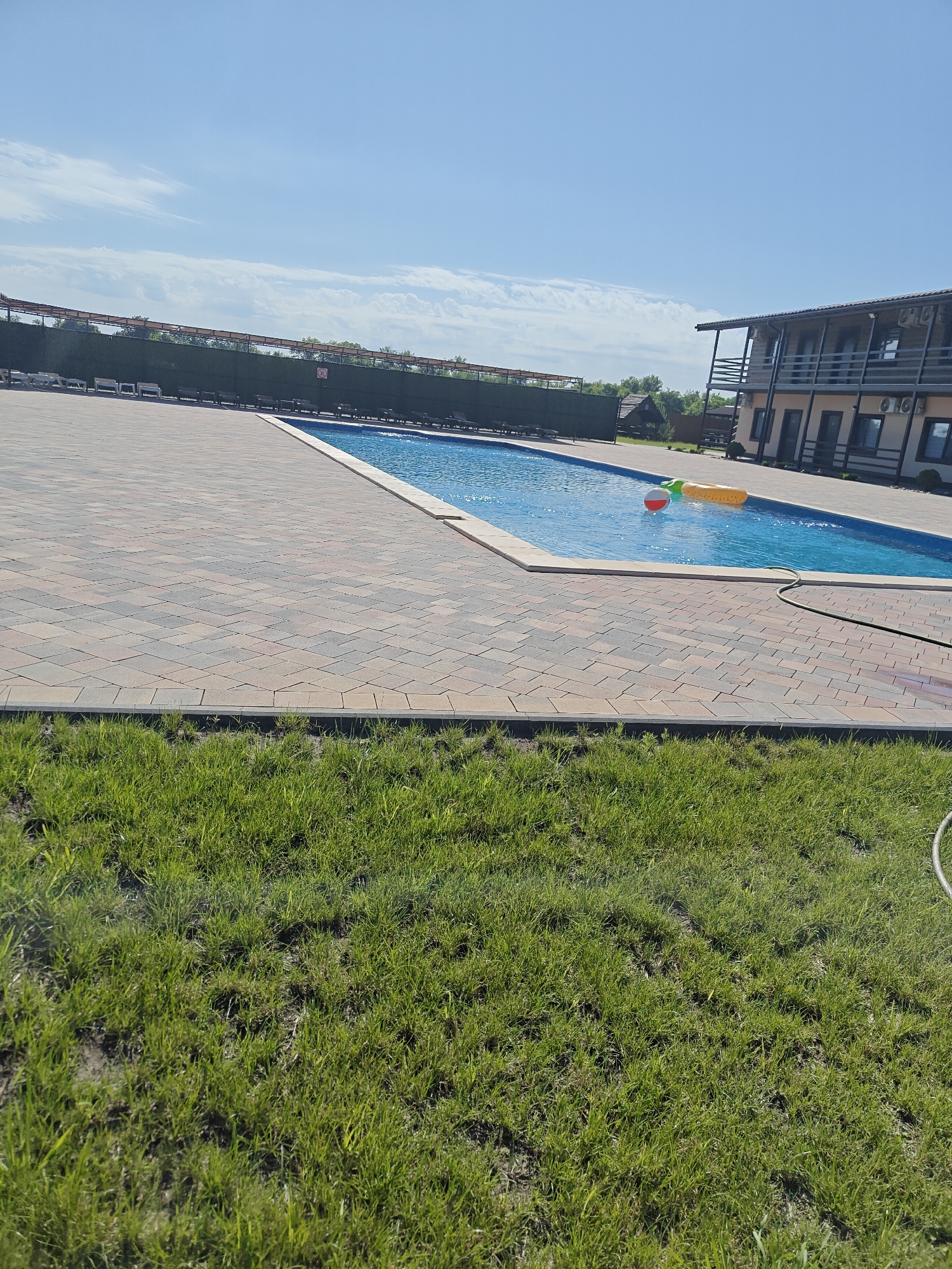The image depicts an outdoor swimming pool area on a sunny day in Shkurupiivka, Ukraine. The pool, filled with clear blue water, is surrounded by a paved patio made of interlocking bricks in shades of brown and gray. In the pool, a yellow inflatable ring and a red and white ball are visible.
To the left of the pool, a dark green hedge forms a boundary, behind which rows of lounge chairs are arranged. Further in the background, trees and a small building with a wooden roof are visible.
To the right of the pool, a two-story building with balconies, windows, and air conditioning units is situated. The building appears to be a hotel or guesthouse, with multiple rooms visible on both floors. The sky above is a clear blue with scattered white clouds. In the foreground, a patch of green grass is present. A green garden hose is visible near the edge of the pool.