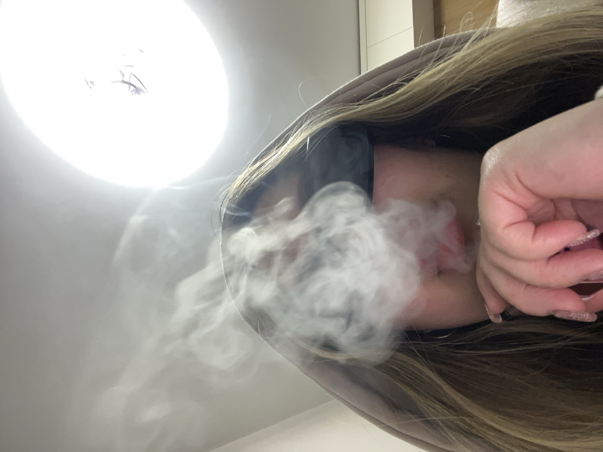 An image, captured from a low-angle, looks upward at a person exhaling a large plume of white smoke or vapor. The subject, likely female, has long, light brown or blonde hair partially covered by a light brown hooded garment. Her face is mostly obscured by the emitted smoke and what appears to be dark sunglasses or a mask. A manicured hand with long, glittery, light pink or clear artificial nails is visible near her mouth, holding something, though the object itself is not clear.
A very bright, circular light source dominates the upper left portion of the frame, casting a strong illumination that creates reflections and slight shadows on a light-colored ceiling. The overall setting appears to be indoors, with artificial lighting being the primary source. The atmosphere is somewhat obscured by the dense smoke. There is no visible text in the image. The scene takes place in Krasnodar, Russia.