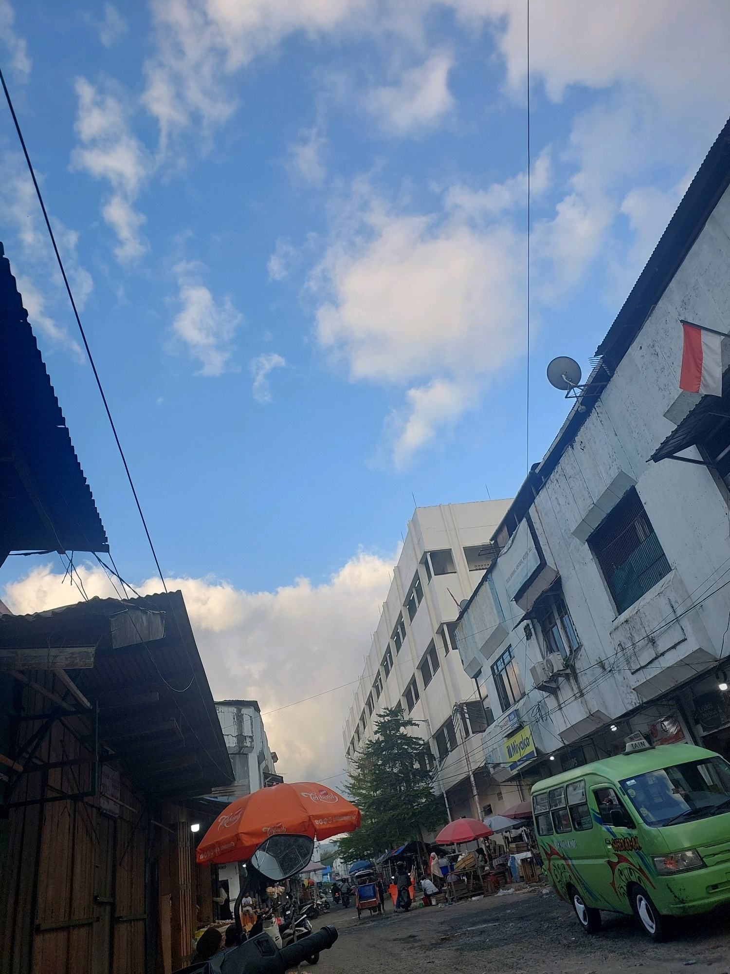 This is a street-level photograph of a busy marketplace in Ambon, Indonesia. The image is dominated by a bright blue sky with scattered white clouds. On the right side of the frame, a multi-story building with a white facade and several windows is visible. A red and white flag, likely the Indonesian flag, is hanging from the upper right corner of this building. In front of the building, a green minibus with decorative patterns is parked on a dusty, unpaved street. The minibus has "TAXI" written on its roof.
To the left of the street, there are market stalls with corrugated metal roofs. One stall is particularly prominent, with wooden walls and a dark, sloped roof. In the foreground, an orange umbrella is open, mounted on what appears to be a motorcycle. Beyond the umbrella, more market stalls, people, and some motorbikes are visible, suggesting a bustling commercial environment. The overall scene depicts a typical day in an urban Indonesian marketplace.