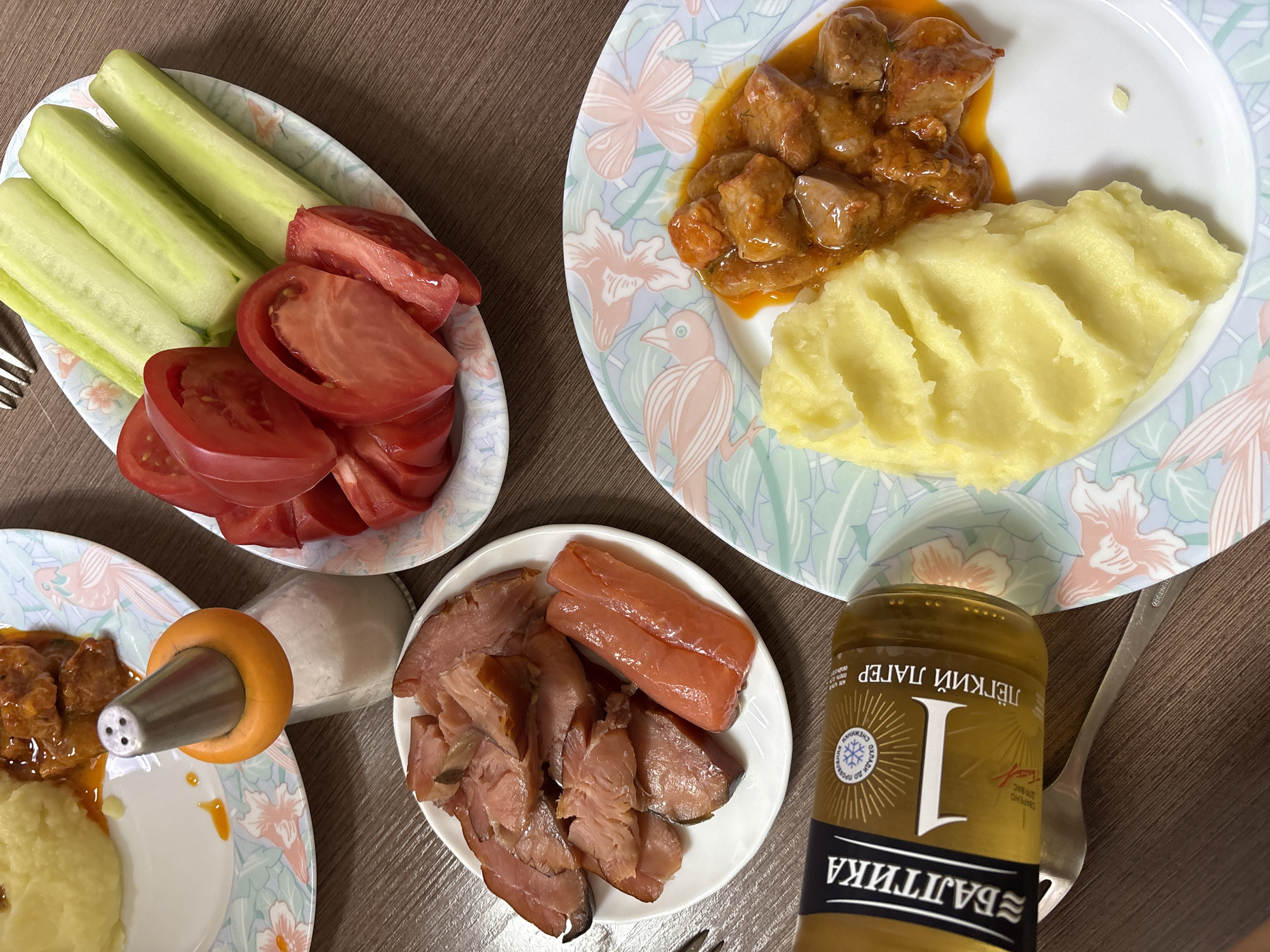 This is a close-up, high-angle photograph of a table laid out with food, likely in a residential setting in Ust-Ilimsk, Russia. The food is arranged on plates with a pastel floral and bird pattern. In the foreground, a bottle of "Baltika 1" lager lies on its side, next to a fork. To the right of the bottle is a plate with a portion of mashed potatoes and a serving of meat stew in a reddish-orange sauce.
Above and to the left of the bottle is another plate containing sliced smoked meats, including what appears to be smoked salmon or trout, and some sausage links. Further up and to the left, a portion of mashed potatoes and meat stew is visible on a plate.
In the upper right quadrant of the image, a plate holds sliced tomatoes and peeled cucumber sticks. Next to this plate, a salt shaker with an orange rim and a metal top is positioned over a small pile of salt. The table surface is a dark brown wood grain. The lighting is diffuse, suggesting indoor illumination. There are no people visible in the image, and the overall impression is of a meal prepared for at least two individuals.