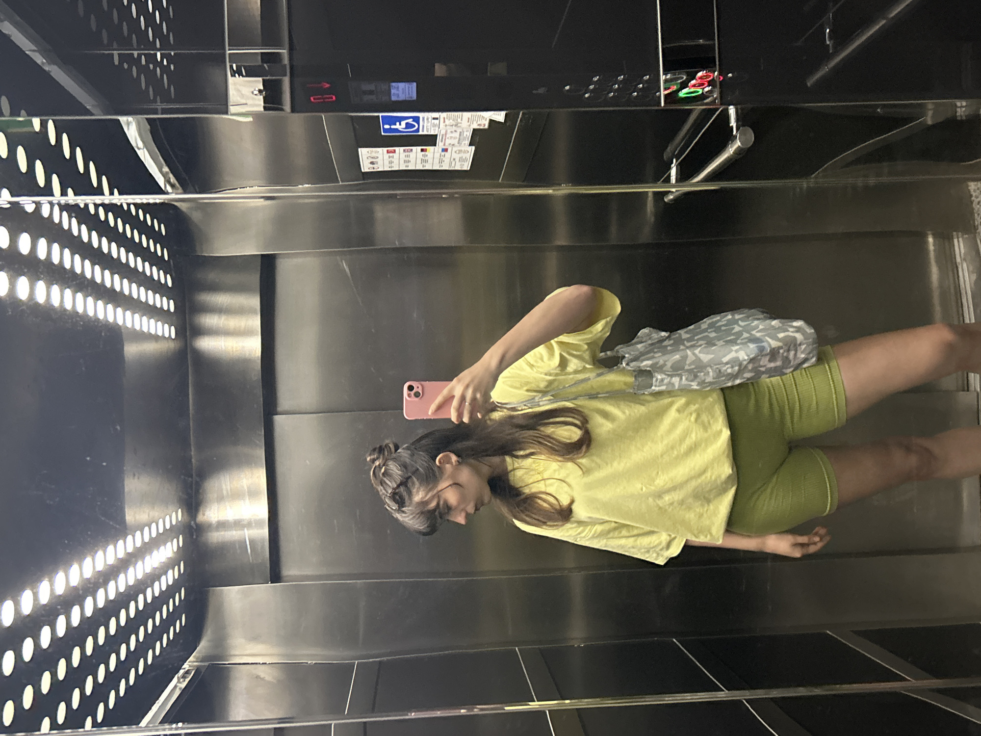 A young woman with long brown hair and wearing a yellow t-shirt and green shorts is taking a selfie in an elevator. She is holding a pink smartphone in her right hand and looking at its screen. She has a grey patterned bag over her shoulder. The elevator interior is stainless steel with a perforated panel on one side. Elevator control panels with buttons and Braille are visible. The location is Alanya, Turkey, based on the context provided.