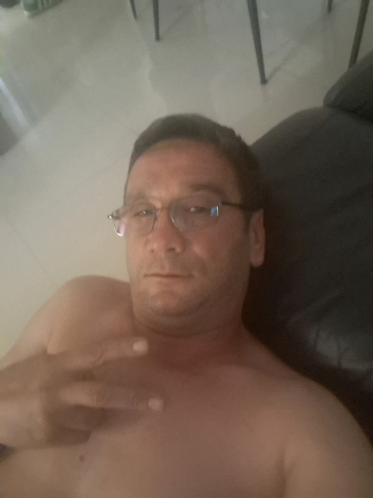 The image shows a man wearing glasses, lying down. His chest and shoulders are visible. He is holding up his left hand, with his fingers extended and in the foreground of the shot. The man appears to be indoors, with a light-colored floor visible in the background, and a dark surface that could be furniture or upholstery to his right. There are also hints of dark, vertical objects in the background, possibly chair legs. The lighting is somewhat dim. The location is the city of Yerocham, Israel.