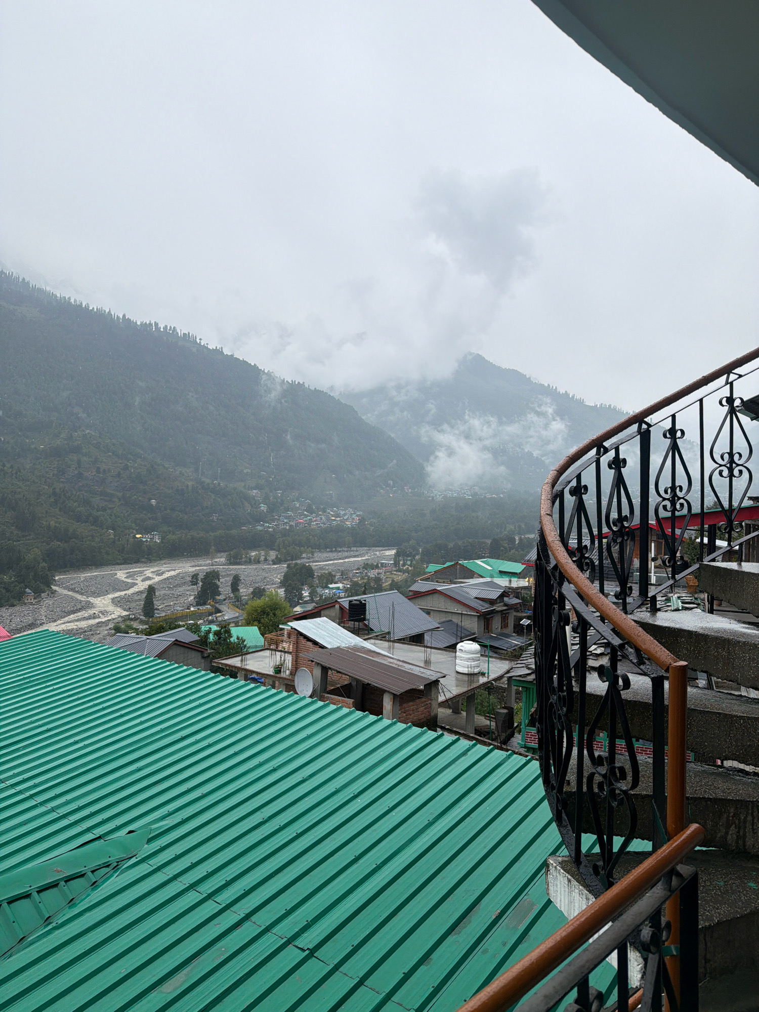 This is a high-angle, outdoor, medium shot of a mountainous landscape in Bahang, India, on an overcast day. In the foreground, a spiral staircase with a black wrought-iron railing and wooden handrails descends from the right. To the left of the staircase, a bright green corrugated metal roof dominates the lower portion of the frame. Beyond the roof, a village or town is nestled in the valley. Numerous buildings with various roofing materials, including gray shingles, red tiles, and more green corrugated metal, are visible. A river with rocky banks meanders through the valley floor. The background is a steep, heavily forested mountain slope that recedes into the distance and is partially obscured by clouds and mist. The overall atmosphere is damp and cool. There are no people or animals visible in the frame.