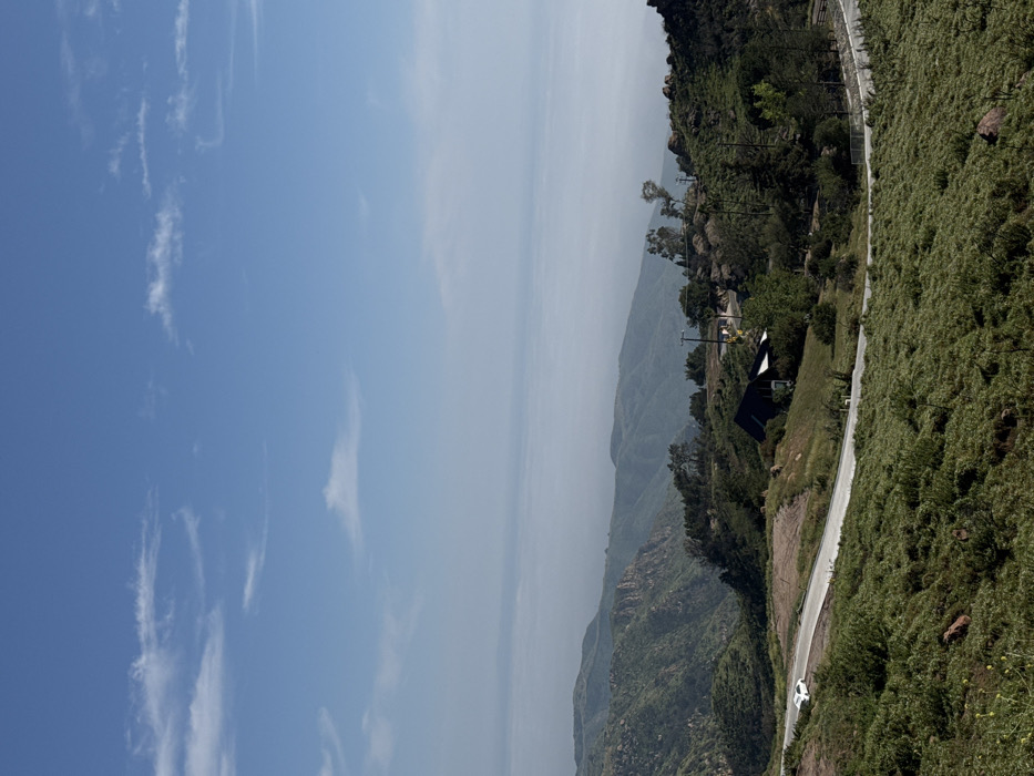 An elevated, wide-angle photograph depicts a mountainous landscape in the Unincorporated Santa Monica Mountains, United States of America, under clear daylight conditions. The sky occupies the upper third of the frame, displaying a light blue hue with sparse, wispy white clouds. The horizon line, marked by atmospheric haze, reveals distant mountains and what appears to be the Pacific Ocean.
In the mid-ground, a paved two-lane road, marked with white lines, curves from the lower-left to the center-right. A single white passenger vehicle, appearing to be a sedan or compact SUV, is visible on the road in the mid-left, oriented towards the right. To the right of the road, a dark-colored building with a sloped, angular roof is situated on a hillside, partially obscured by trees.
The terrain is characterized by rolling hills and steep slopes covered predominantly in green, low-lying brush and scattered trees. Exposed reddish-brown rocks are visible throughout the landscape, particularly on the lower left and in the vicinity of the building. Several utility poles with power lines traverse the hillsides. No individuals are visible in the scene.