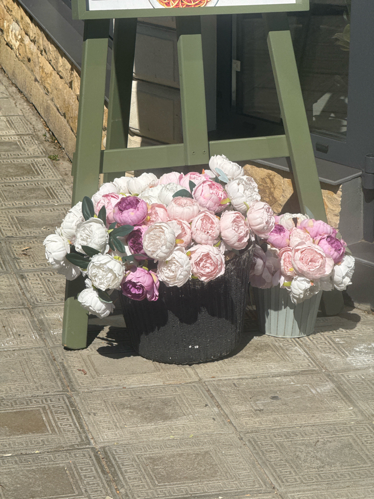 A still life of artificial flowers on a cobblestone sidewalk in front of a building with a stone facade. Two pots of artificial peonies, one large black pot and a smaller pale blue striped pot, are placed on the sidewalk. The flowers are a mix of white, light pink, and deep pink/purple colors. A green easel-like structure, likely holding a sign that is not visible, stands behind the pots. The sunlight casts shadows on the ground. The scene is set in Anapa, Russia.