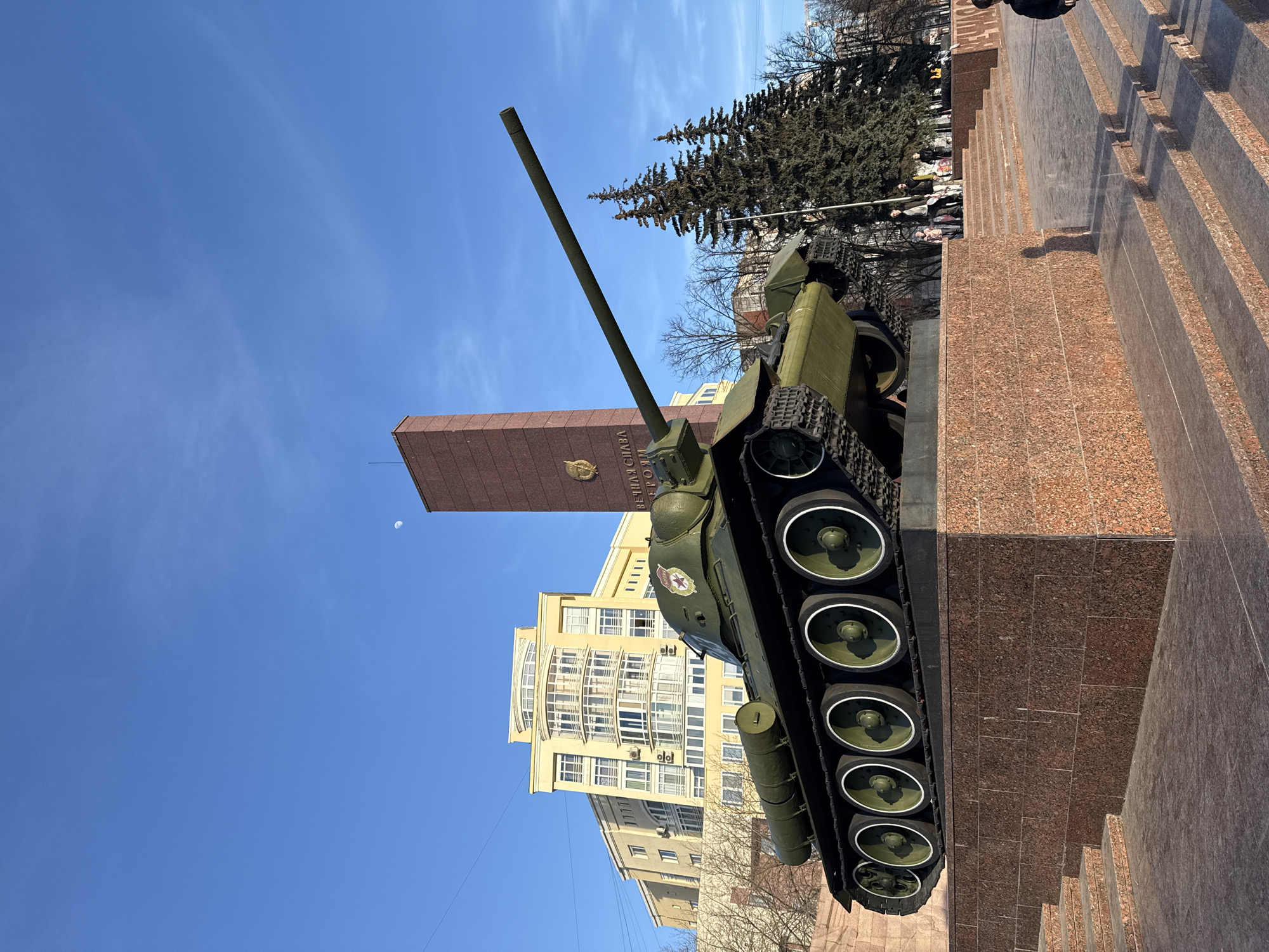 The image depicts the T-34-85 tank monument located on Komsomolsky Prospekt, Perm, Russia. The primary subject is an olive-green T-34-85 tank, mounted on a multi-tiered pedestal constructed of polished red granite. The tank is positioned facing right, with its main gun barrel extended horizontally towards the right. A red star emblem with a gold hammer and sickle is visible on the tank's turret.
Behind and to the right of the tank, a tall, rectangular stele monument, also made of reddish-brown material, rises against the sky. Inscriptions in Cyrillic, reading "ВЕЧНАЯ СЛАВА ГЕРОЯМ" (Eternal Glory to the Heroes), are visible on its face, along with an emblem. To the right of the stele, a cluster of dark green coniferous trees stands tall. In the background on the left, a large, multi-story yellow building with numerous windows and balconies is visible. Bare deciduous trees are situated in front of this building.
The foreground consists of polished red granite steps leading up to the tank's pedestal. Scattered individuals are present in the mid-ground and background, walking near the monument and trees on the right side of the frame; their features are indistinct. A waxing crescent moon is visible in the clear, bright blue sky in the upper center-right portion of the image. The scene is illuminated by direct sunlight, casting shadows.