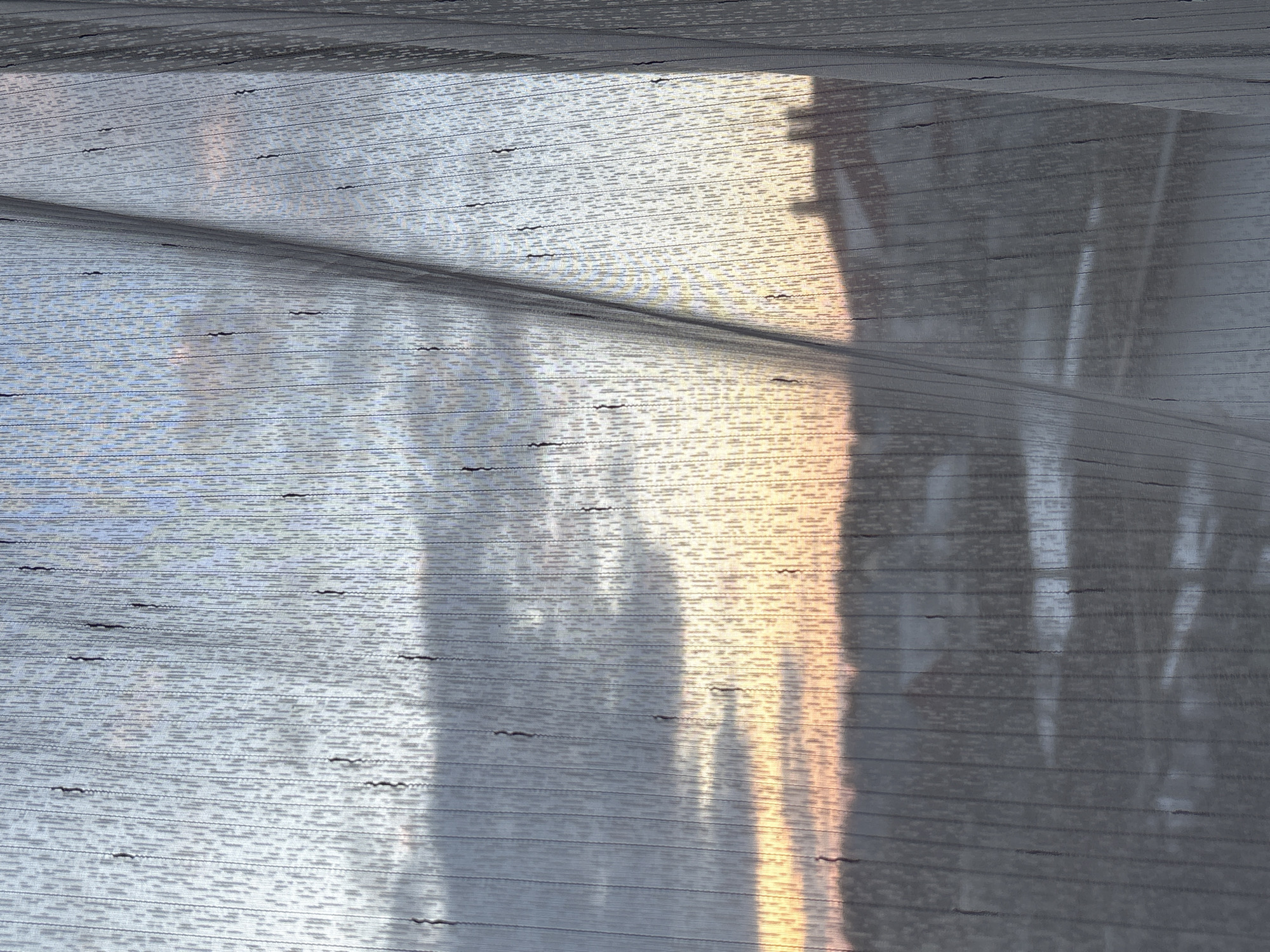 The image shows an abstract close-up of a textured surface with light and shadow patterns. The surface appears to be a woven material with horizontal lines and small dark markings. The light creates variations in color, ranging from light blue-gray to warm golden tones. Shadows suggest the presence of objects or structures casting their shapes onto the material. The location is Прикамский, Russia. The image doesn't depict people, actions, or interactions.