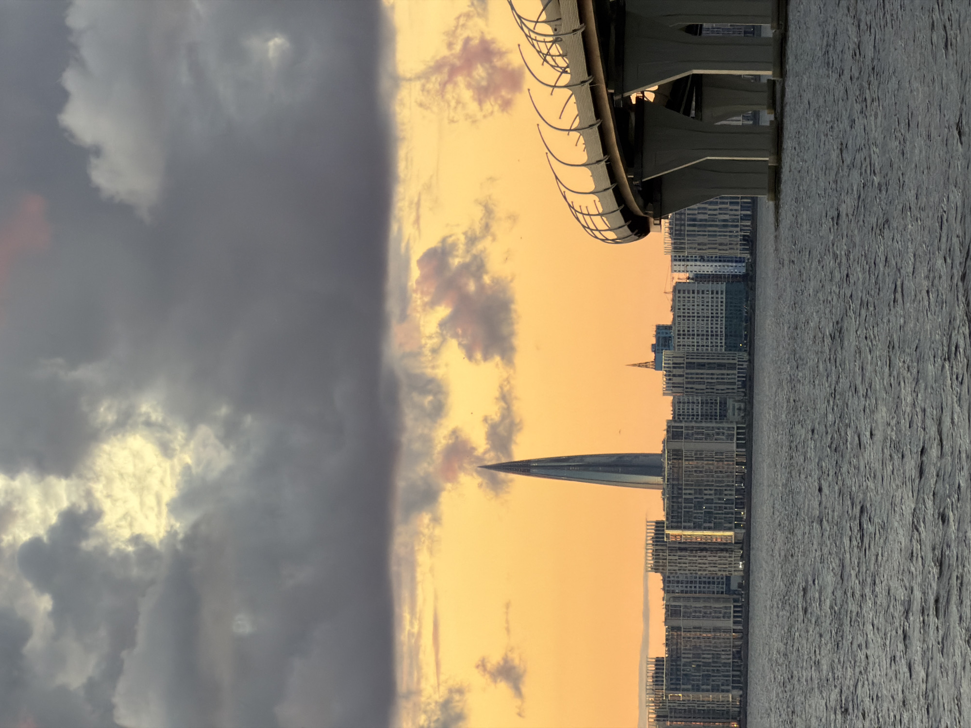 The image shows a view of the Gulf of Finland with a modern cityscape and a bridge in the foreground. On the right side of the image, a large elevated highway bridge curves into the frame, supported by multiple concrete pillars extending into the water. The bridge has a metallic railing system.
In the background, the skyline of Saint Petersburg, Russia, is visible across the water. Dominating the center of the cityscape is the Lakhta Center, a striking modern skyscraper with a distinctive pointed spire. To the left and right of the Lakhta Center are numerous residential and commercial buildings, characterized by their uniform height and repetitive architectural design, suggesting a newly developed area. The water in the foreground is rippled and grey, reflecting the overcast sky.
The sky is a dramatic display of clouds. Dark, heavy clouds occupy the upper portion of the image, with a bright, yellowish-orange glow breaking through in the center, indicating either sunset or sunrise. Scattered lighter clouds, tinged with pink, are also present against the vibrant sky. The overall lighting suggests a transitional time of day, with dramatic cloud cover. There are no people or specific actions depicted in the scene. The overall atmosphere is one of urban development and natural beauty combined.