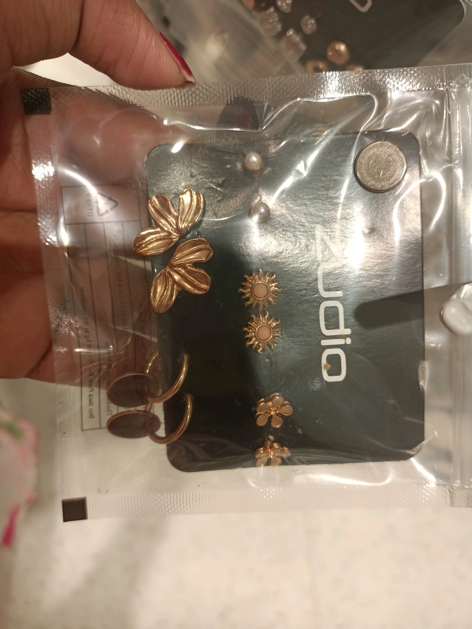 The image shows a close-up of a set of earrings in plastic packaging. The earrings are gold-toned and include various designs such as butterflies, suns, and small flowers. The packaging is clear plastic with a white backing card that has some text and symbols printed on it, including a recycling symbol. A hand with red-painted fingernails is holding the packaging from the top left corner. The image does not provide any information about the city of Barrackpore, India, nor does it depict any outdoor environment. The focus is solely on the product within the packaging.