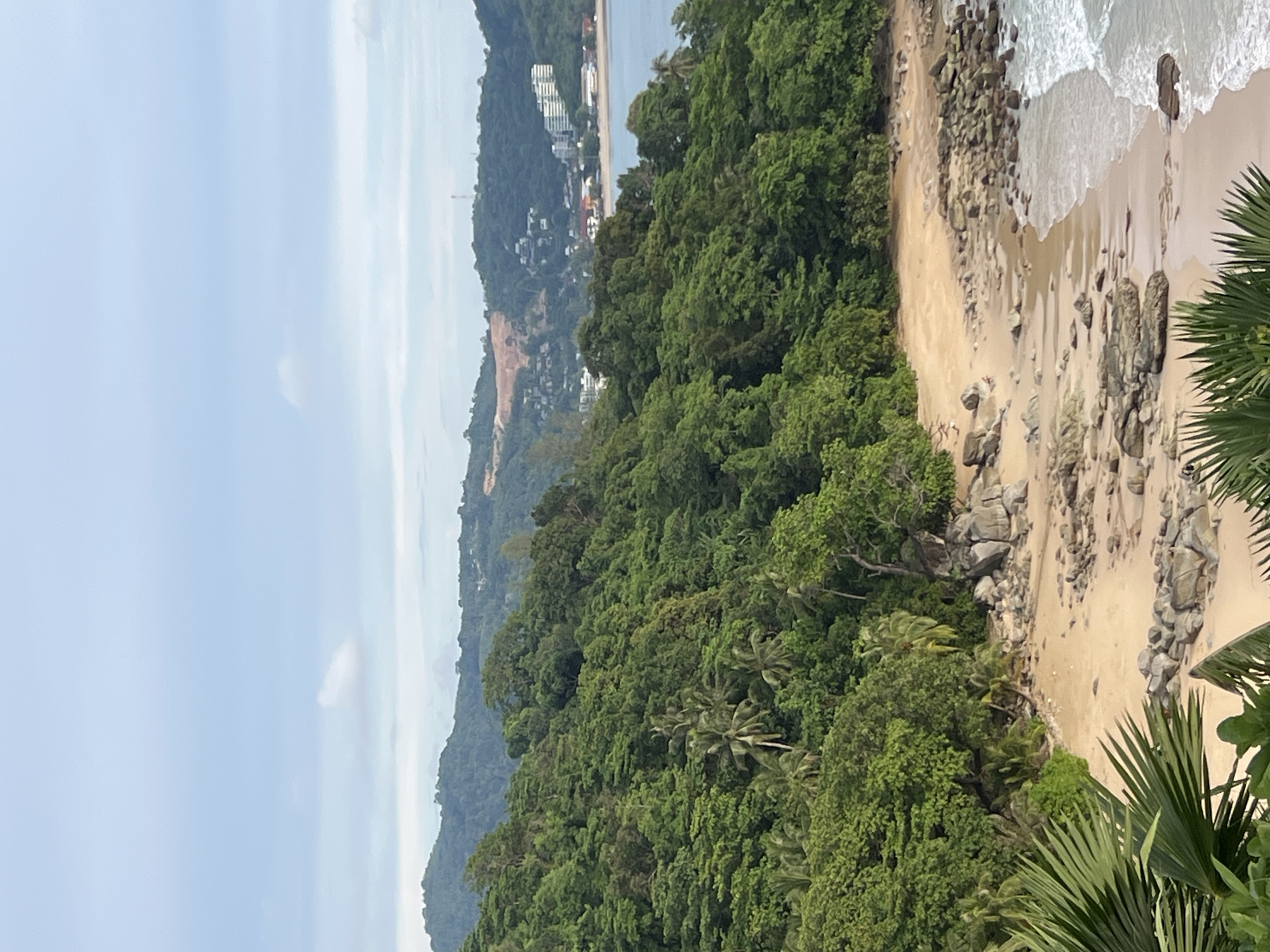 The image captures a scenic view of a tropical coastline in Ban Bang Thao, Thailand. In the foreground, lush green foliage, including palm fronds, frames the scene. A sandy beach stretches along the coastline, dotted with numerous grey rocks of varying sizes. Gentle waves lap at the shore, creating a serene atmosphere.
Beyond the beach, a dense, verdant jungle covers a steep hillside, with palm trees interspersed among the other trees. In the distance, a town or resort area is visible along the coast, with buildings nestled amongst the hills and a larger building with multiple floors prominently featured. The background consists of rolling hills and a hazy sky with scattered clouds. The overall impression is one of natural beauty and coastal development.