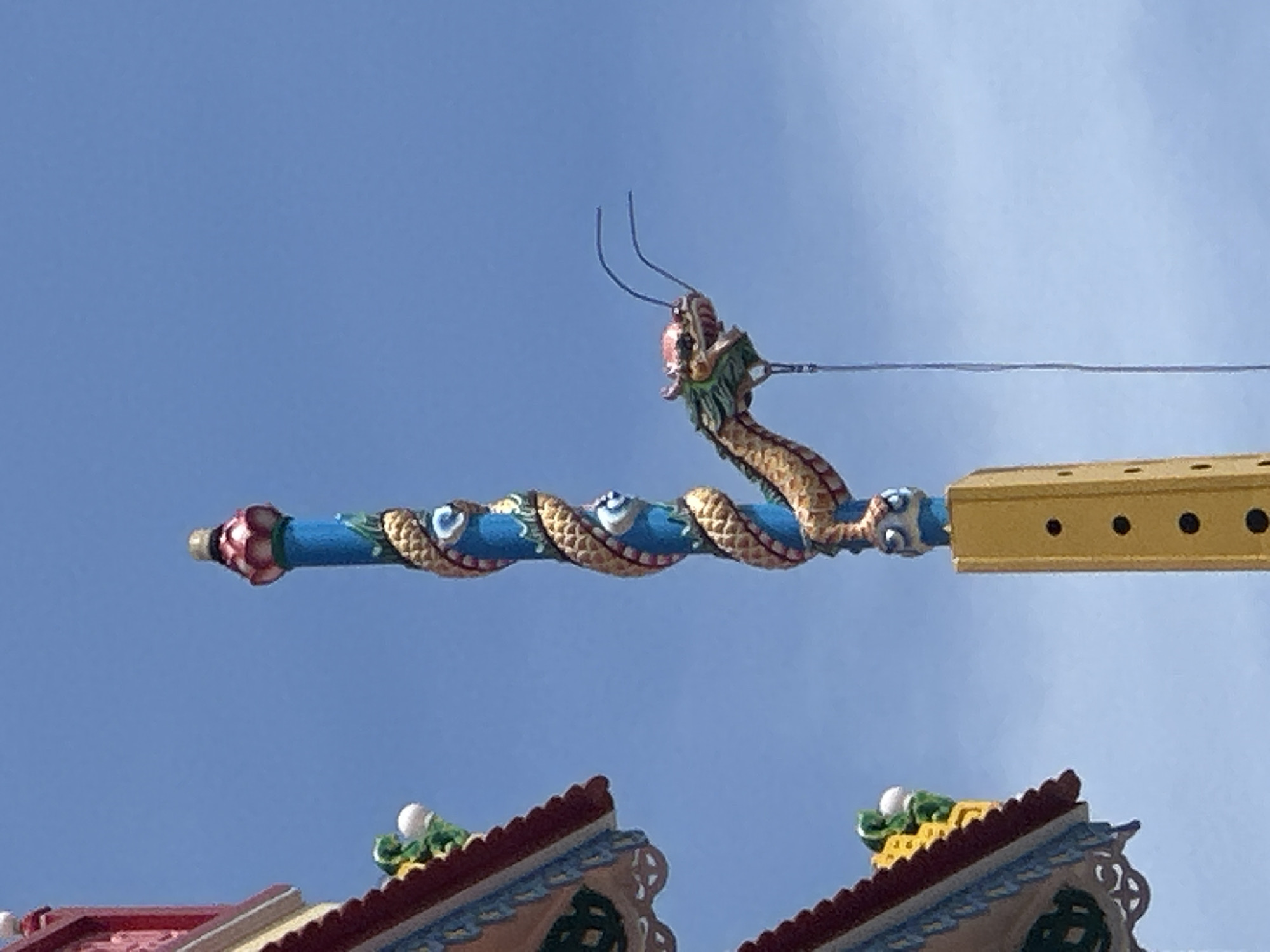 A bright blue sky serves as the backdrop for a decorative pole adorned with a golden dragon sculpture. The dragon coils around the blue pole, its head facing towards the right, with red accents on its face and green accents around its eyes. The pole itself is topped with a pink and red lotus-like finial and features blue and white circular motifs interspersed with the dragon's scales. Below the dragon, a yellow rectangular structure with black circular cutouts is visible. To the left of the pole, a portion of a building's roof is visible, featuring red and yellow tiled sections with decorative green and white elements. The scene appears to be outdoors, likely in an area with traditional architecture, possibly a temple or cultural site in Nha Trang Ward, Vietnam. There are no people or animals depicted in the image.