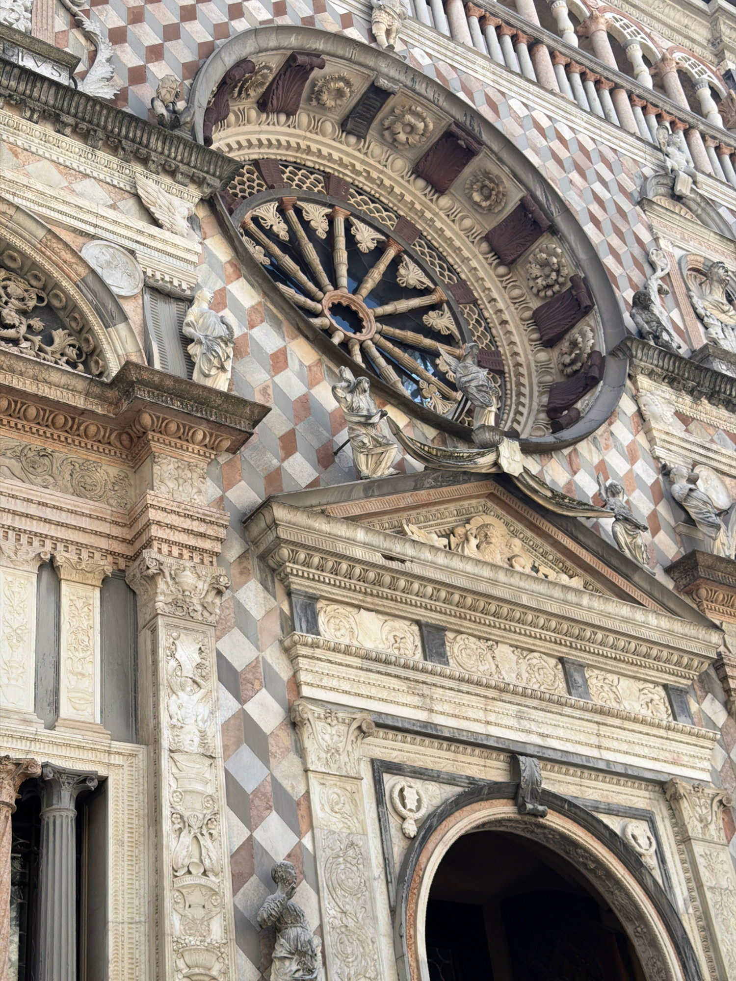 This is a close-up, low-angle shot of the facade of a building, likely a cathedral or a historical monument, characterized by intricate architectural details. The building features a repeating diamond pattern in shades of pinkish-brown, cream, and grey, creating a visually striking mosaic effect. Above this pattern, a large, ornate rose window is visible, its intricate stone tracery forming a radial design. The window appears to be made of dark wood or metal spokes against a darker background, possibly glass.
Below the rose window, the facade is adorned with multiple layers of carved stone. These carvings include decorative moldings, friezes with figural reliefs, and classical architectural elements like columns and pediments. Several small statues of figures, likely religious in nature, are placed on ledges and at architectural junctures. One statue on the lower left appears to be a winged figure. Another, on the lower right, is a standing female figure. The architecture suggests a Gothic or Renaissance style.
The lighting suggests it is daytime, with soft light illuminating the details without harsh shadows, possibly indicating an overcast day or late afternoon light. The stone surfaces show signs of age and weathering, contributing to the historical feel of the structure. The scene is static, focusing entirely on the architectural grandeur of the building. No people are clearly identifiable as active participants in a scene, only the sculpted figures. The location context points to Mozzo, Italy. No visible text is discernible in the image.