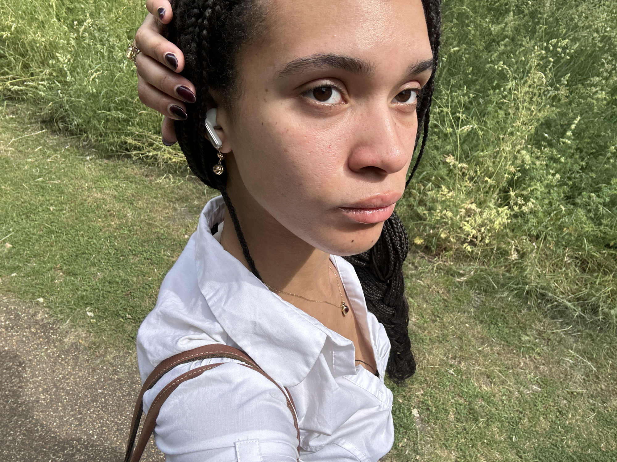 A young woman with braided hair is centered in a medium shot, positioned outdoors in a park environment. She is wearing a white collared shirt and a gold necklace with a red pendant, with the brown strap of a bag visible on her shoulder. A white wireless earbud is in her left ear with a small gold charm hanging from it. Her left hand, adorned with a gold ring and dark nail polish, is near her head, with only the back of the hand visible. The background consists of green grass and some wildflowers. To the left, there is a paved area. The location appears to be a park in London, United Kingdom, due to the typical grass texture and flora.