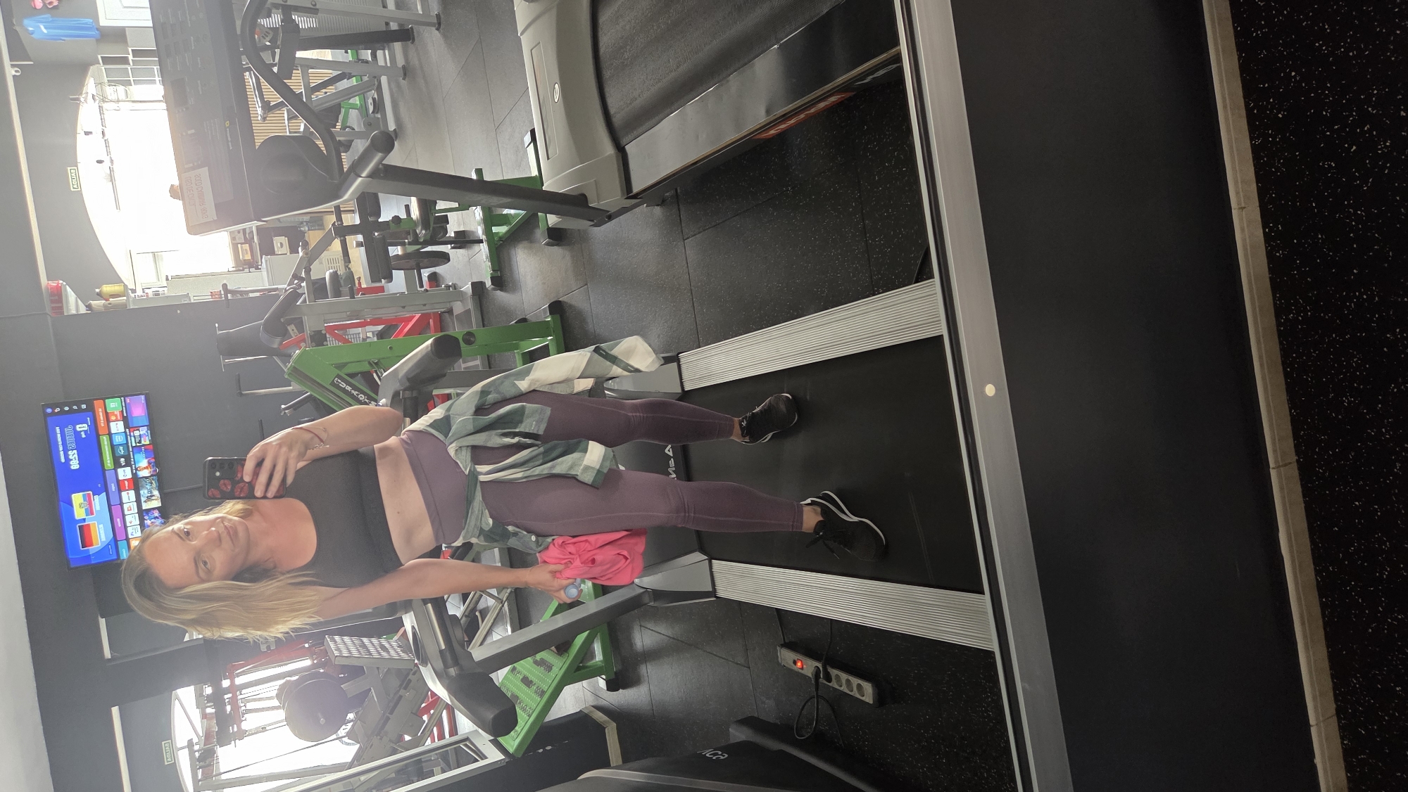 A woman is taking a selfie in a gym. She is wearing a black sports bra, purple leggings, and black sneakers. A green and white plaid shirt is tied around her waist. She is standing on a treadmill. In the background, there are various gym equipment, including weight machines and other treadmills. A television screen is mounted on the wall, displaying a menu with flags of Spain and Germany. The lighting suggests it is daytime. The woman appears relaxed and is looking at her phone, which she is using to take the picture. The gym is well-lit with modern equipment and a dark, speckled floor.