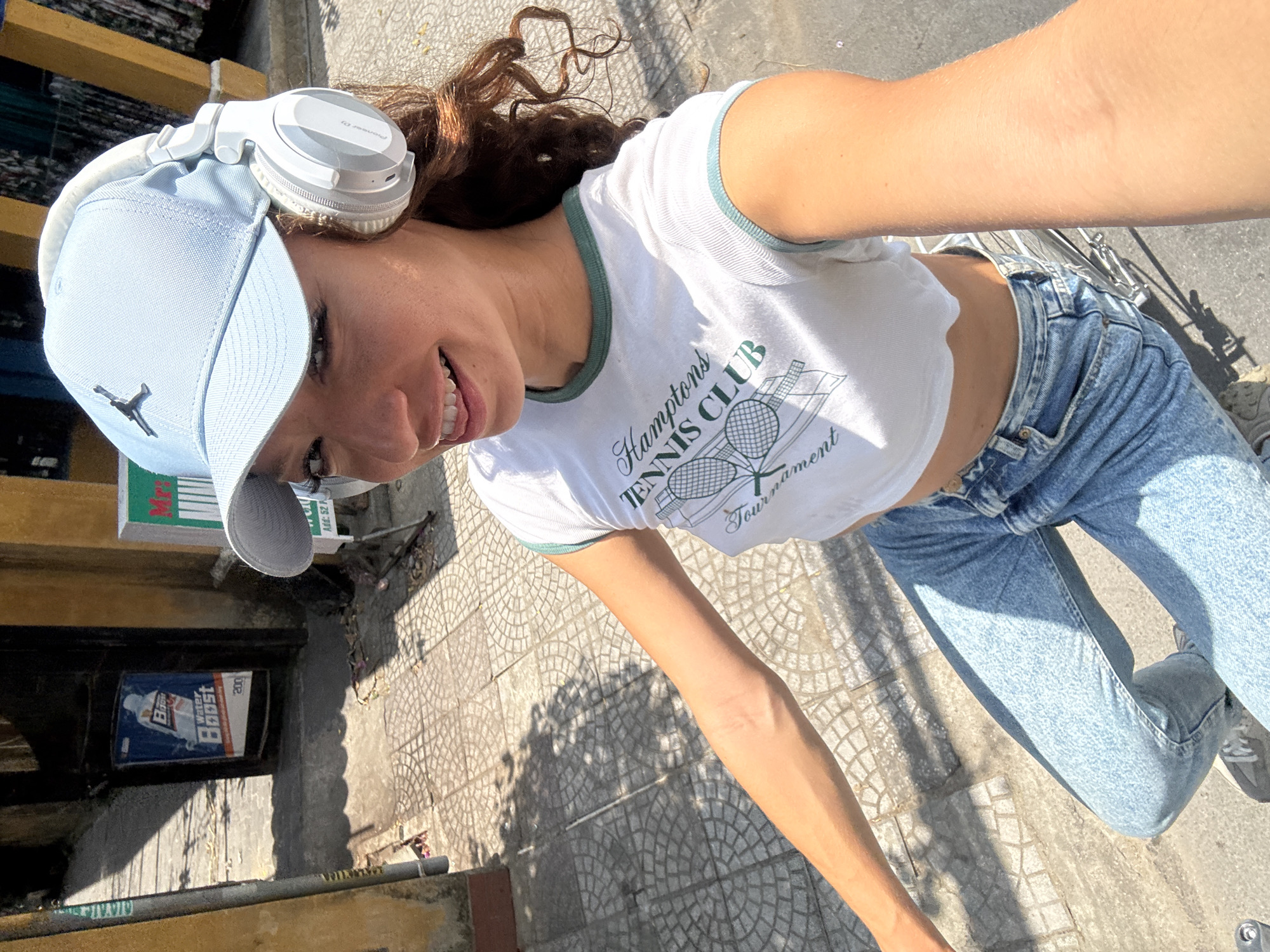 A woman poses for a selfie outdoors on a sunny day in Hội An Ward, Vietnam. She is wearing a light blue baseball cap with a "Jordan" logo, white headphones, a white crop top with "Hamptons TENNIS CLUB" printed on it, and light-wash blue jeans. She is smiling and looking towards the left. The ground is tiled with a geometric pattern. In the background, there is a storefront with various items and a "Water Boost" advertisement. The setting appears to be an urban street.
