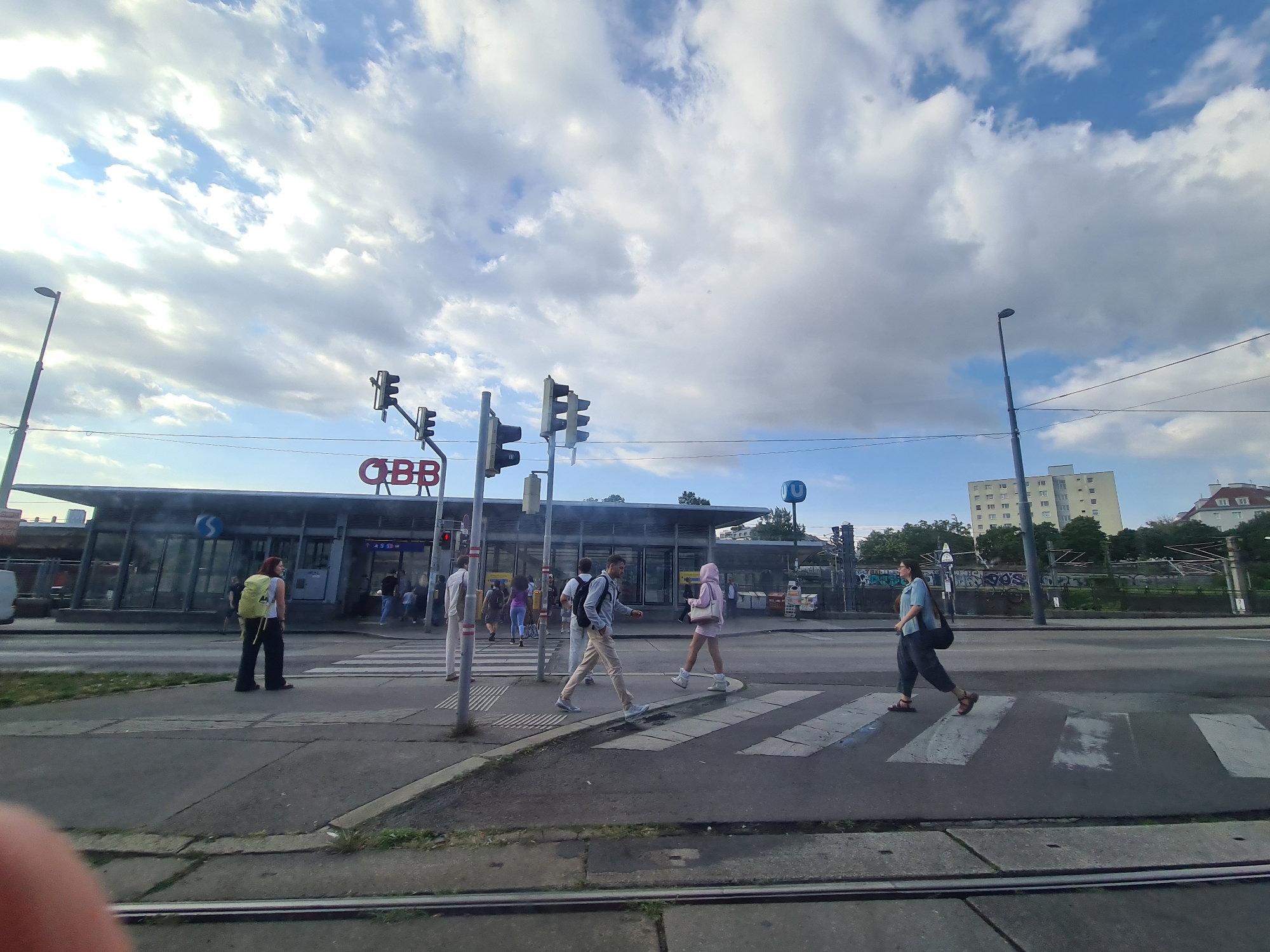 This image captures a street scene in Vienna, Austria, likely near a train station, given the prominent "ÖBB" (Austrian Federal Railways) signage on the building in the background. The weather appears partly cloudy, with a mix of blue sky and white clouds. Several pedestrians are visible on the sidewalk and crossing a zebra crossing.
In the foreground, a person with a bright yellow backpack is standing on the sidewalk, facing away from the camera. To their left, several other individuals are seen near the entrance of the building. Further to the right, a group of people are crossing the street. One individual, wearing a light-colored hoodie and shorts, is walking across the zebra crossing, as is a person in a light blue shirt and dark pants carrying a shoulder bag. Another person, dressed in white pants and a light-colored shirt, is standing near a traffic light post on the right side of the image.
The building in the background is a modern structure with a glass facade and a prominent red "ÖBB" logo. Traffic lights and lampposts are also present along the street. The scene suggests a busy urban environment, possibly a transportation hub. The presence of a blue "U" symbol, indicative of the Vienna U-Bahn (subway) system, can be seen on a pole in the background, further confirming the location within Vienna and suggesting proximity to public transport. The street itself has tram tracks visible in the foreground.