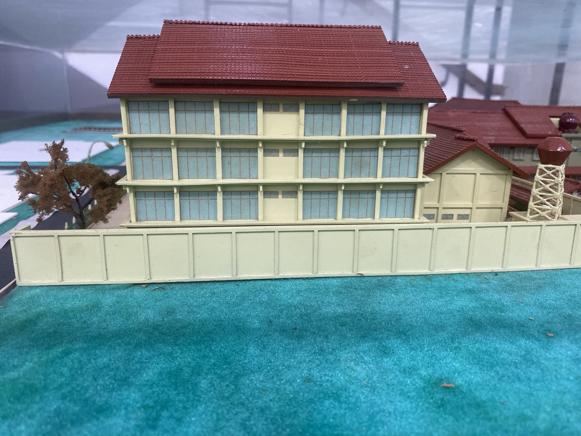 This is a close-up, eye-level, outdoor shot of a model of a building in Banda Aceh, Indonesia. The model features a three-story building with a red tiled roof, light yellow walls, and numerous light blue windows. A light yellow fence runs across the front of the model, and behind it, a small, brown, leafless tree is visible. The model is placed on a flat, teal-colored surface that represents grass or ground cover. In the background, other parts of the model building and a water tower can be seen. The overall scene suggests a diorama or architectural model display.