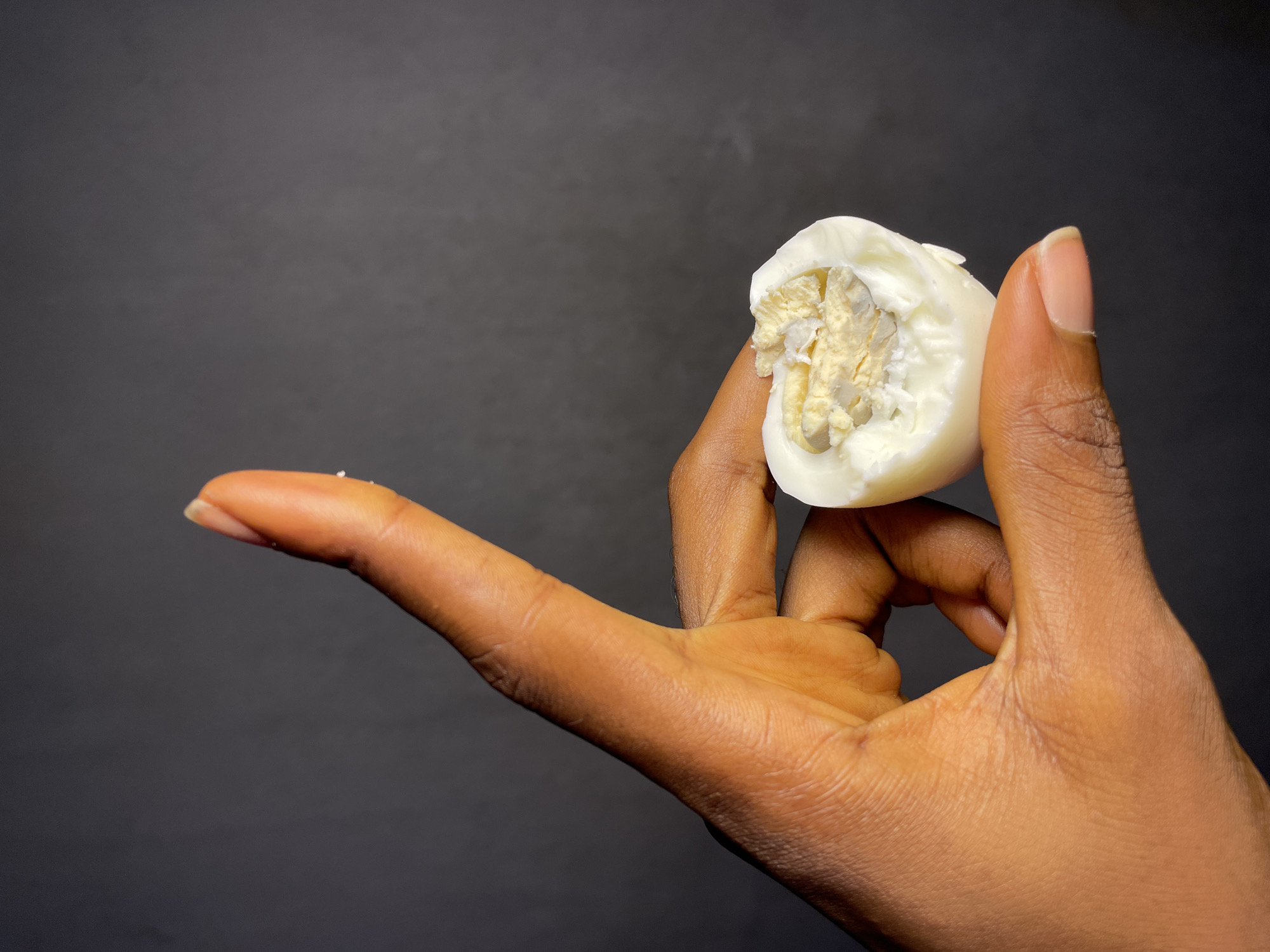 A close-up shot presents a dark-skinned hand holding a halved, hard-boiled egg. The egg's white is smooth and intact, while the yolk appears somewhat crumbled. A small piece of egg white is visible on the extended index finger. The background is a solid, dark gray color. The image was not taken in Nkpor, Nigeria.