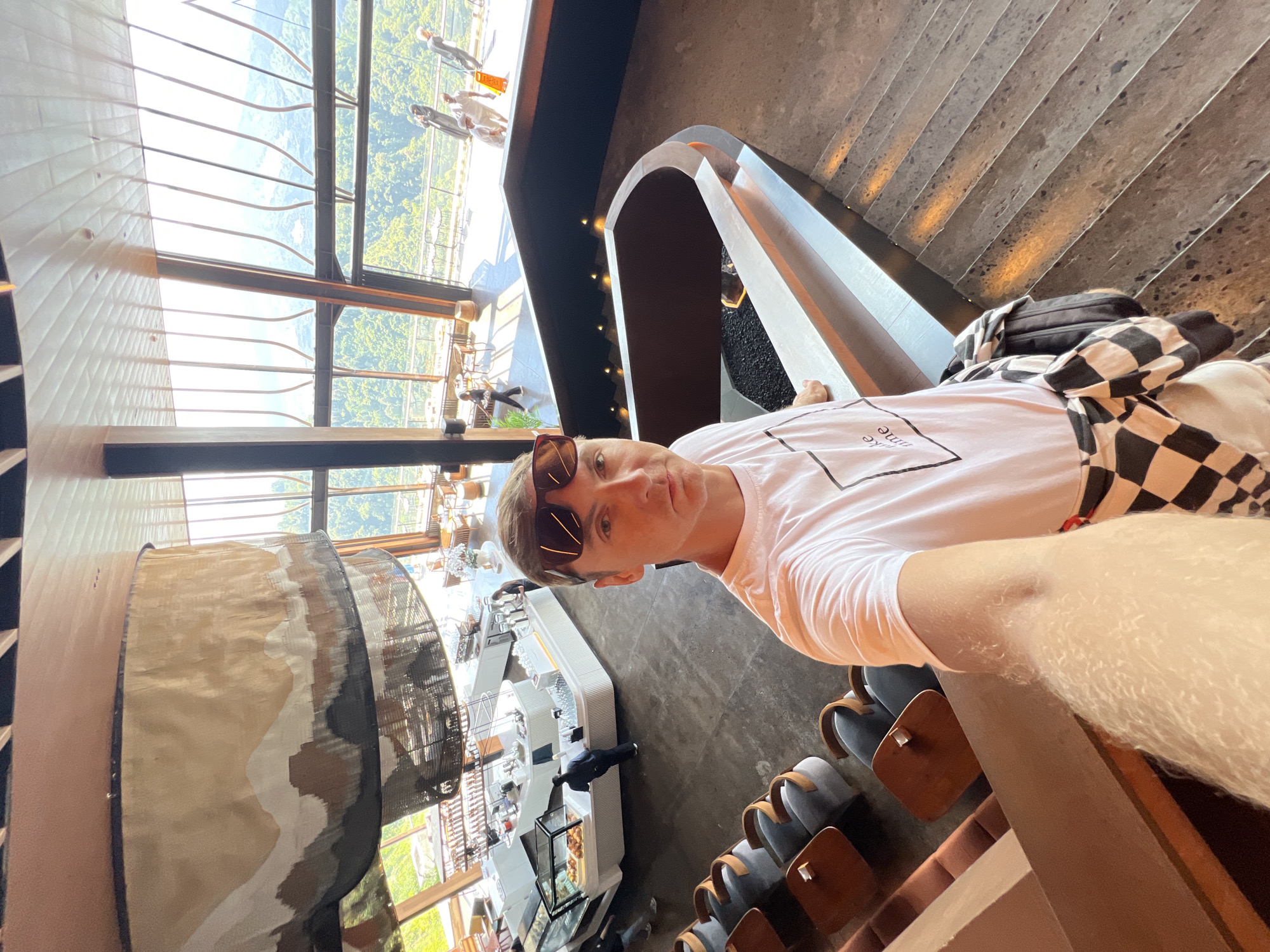 A man wearing sunglasses and a white t-shirt takes a selfie in a modern, multi-level cafe with large windows overlooking a lush, green landscape. The cafe features a curved counter with a display of baked goods, seating areas with chairs and tables, and a staircase with warm accent lighting. The man is standing on the stairs, with his arm extended to take the photo. In the background, other patrons can be seen dining and walking around. The overall ambiance suggests a contemporary and comfortable space designed to maximize the scenic views. The location is identified as a cafe in Kintamani, Indonesia, known for its picturesque setting.