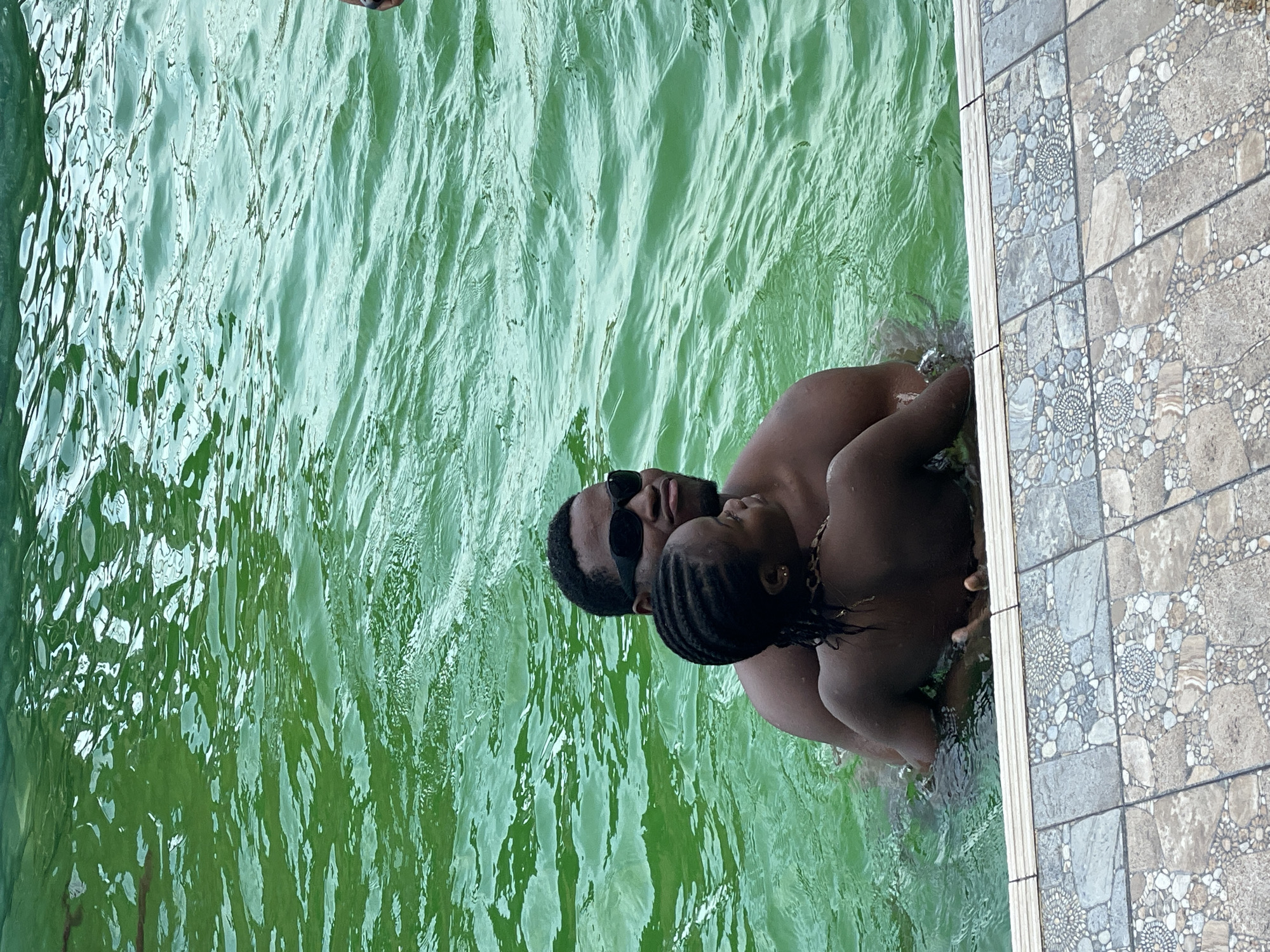 A father and child are in a swimming pool. The father, wearing sunglasses, has his arm around the child, who is looking away from the camera. The water is green, and the edge of the pool is visible in the foreground, tiled with a mosaic pattern.