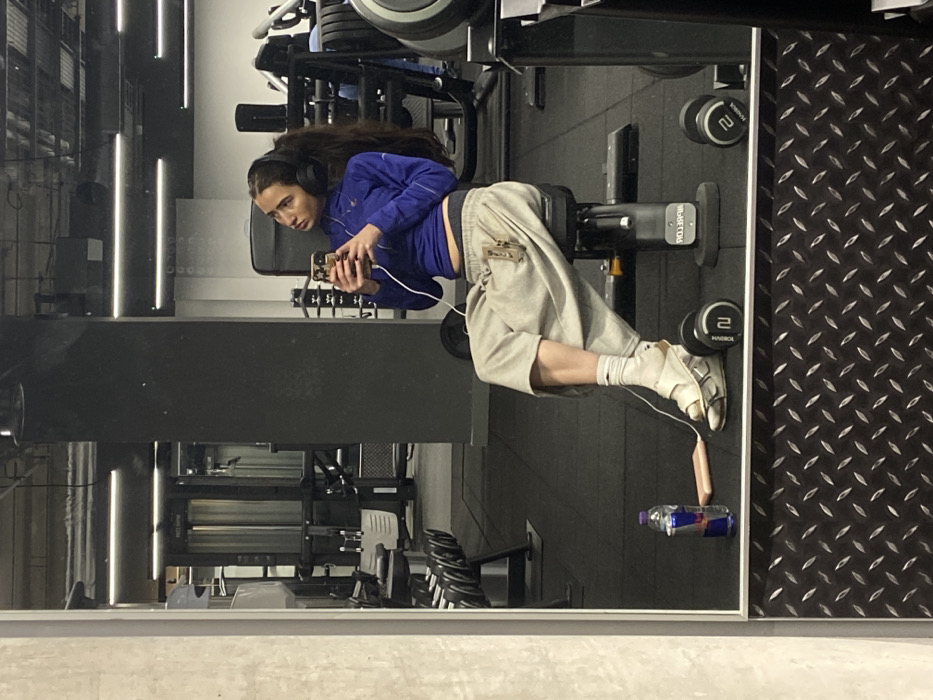A female subject is seated on a black adjustable gym bench, observing her reflection in a large mirror within a gymnasium in London, United Kingdom. She wears a blue long-sleeved athletic top, grey sweatpants, white socks, and white sandals, accessorized with black over-ear headphones. Her right hand holds a smartphone with a light-colored case, connected by a white cable to a light pink/beige power bank resting on the black rubberized floor. Her gaze is directed towards the upper right of the frame. Adjacent to her on the floor are a red and silver Red Bull can and two black dumbbells, each marked "2". The background, visible through the mirror, displays standard gym equipment, including weight machines, dumbbell racks, and dark structural pillars, illuminated by overhead strip lighting. A reflective diamond plate strip borders the bottom of the mirror.