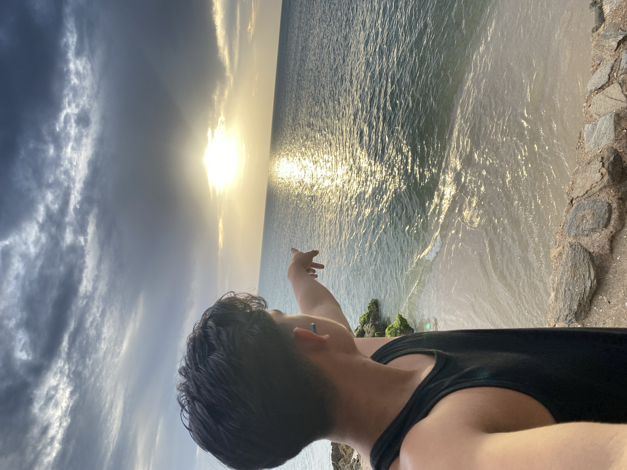 A young male, seen from behind and from the shoulder up, stands on a rocky outcrop overlooking the sea. He wears a black tank top and points his left arm towards the sun setting over the ocean. The sun's rays create a shimmering path across the water's surface. Dark, dramatic clouds fill the upper portion of the sky. The background depicts a serene beach with gentle waves lapping the shore. The scene is set in Juan Griego, Venezuela, known for its stunning sunsets and coastal beauty.