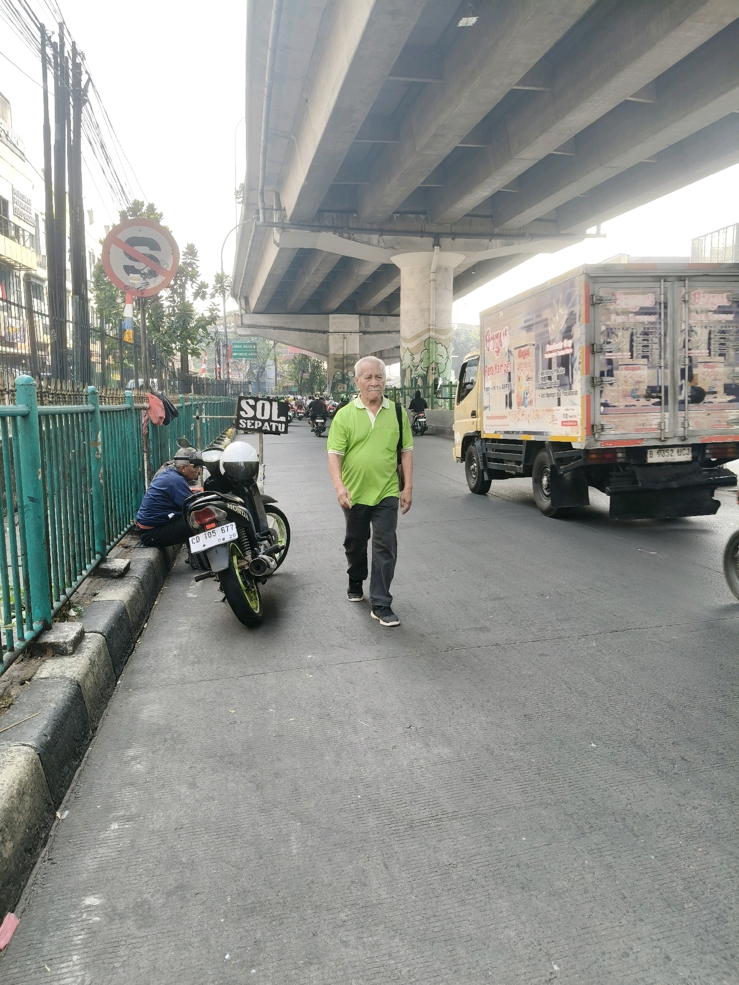 An elderly man with grey hair and a mustache, wearing a green short-sleeved polo shirt and dark pants, walks on a road under a concrete overpass in South Tangerang, Indonesia. A yellow truck with a box trailer is driving on the right side of the road. On the left side of the road, a man wearing a blue jacket and a cap sits on the curb next to a parked motorcycle. A sign with red and white stripes indicates "SOL SEPATU" which translates to "Shoe Shine." The environment appears to be an urban setting with streetlights, buildings, and traffic.