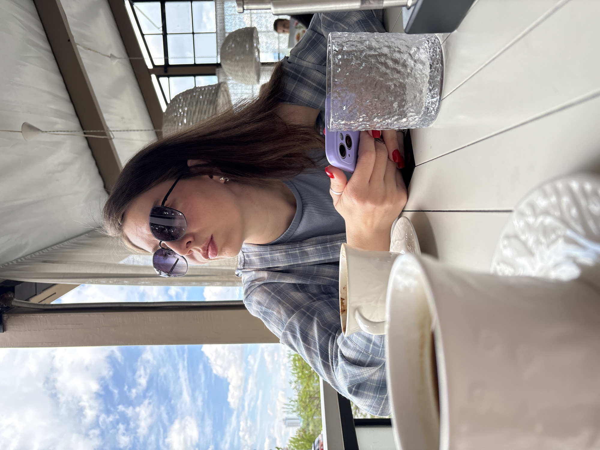 A woman with long brown hair and sunglasses is seated at a white table, holding a purple smartphone. A glass of water and two white mugs are also on the table. The scene is located on an open-air terrace or patio, as evidenced by the partly visible sky, trees, and buildings, which suggest the city of Krasnoyarsk, Russia.