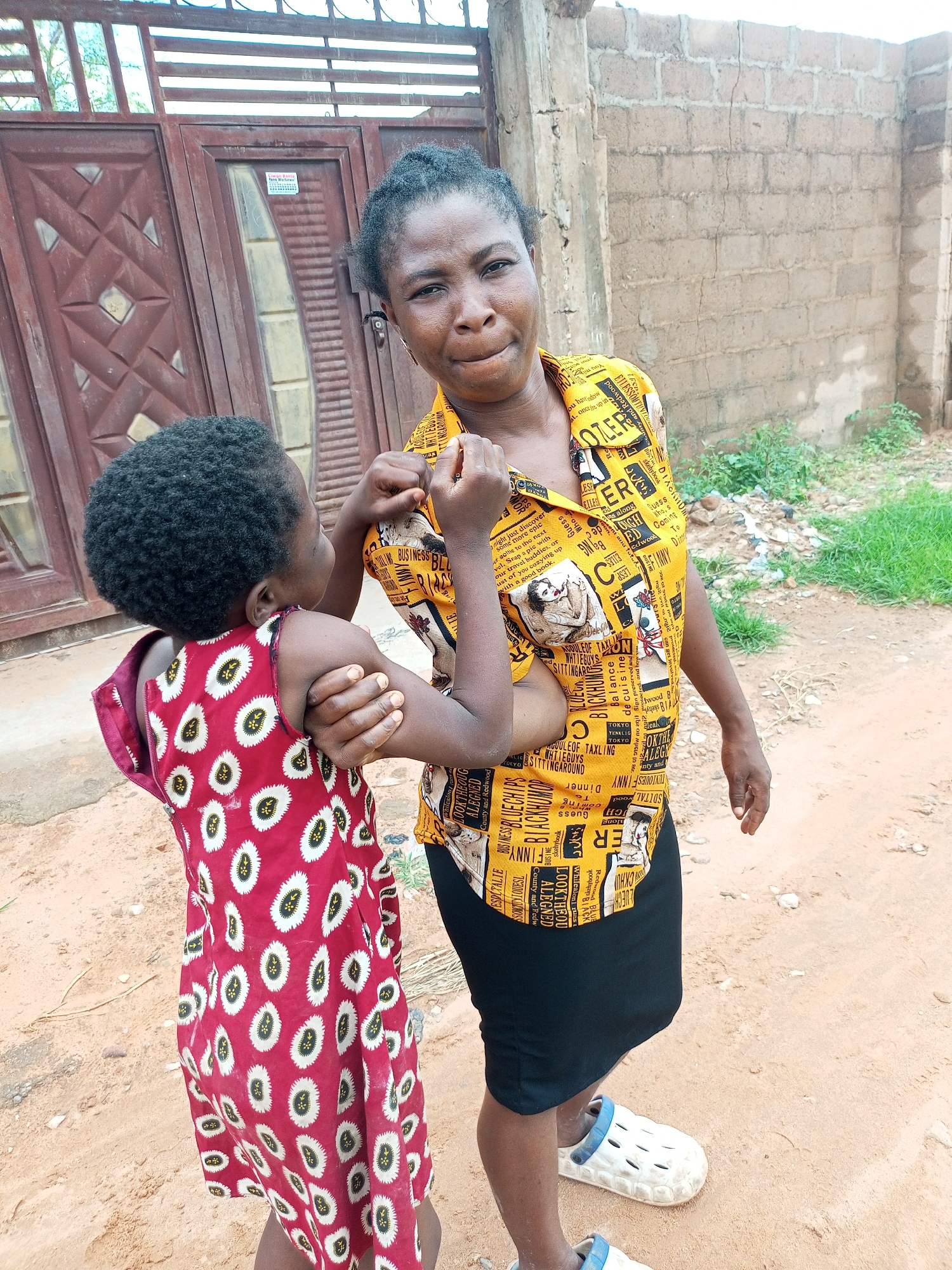 The image depicts an outdoor scene in what appears to be the city of Tempure, Nigeria. In the foreground, a woman with dark hair pulled back is looking slightly off-camera with a neutral expression. She is wearing a yellow patterned shirt with text and images, and a black skirt. She has on white clogs with blue trim. To her left, a young child with dark curly hair is facing away from the camera, embracing the woman's arm. The child is wearing a red sleeveless dress adorned with a repeating pattern of white oval shapes outlined in black with yellow centers.
Behind them, a large, rustic wooden gate with metal bars is visible. The gate is a dark reddish-brown color and appears to be divided into two sections. A portion of a light-colored brick wall forms the background on the right side of the image, with some green vegetation growing at its base. The ground is a sandy, reddish-brown dirt with some small stones and tire tracks visible. The overall setting suggests a residential or semi-developed area.