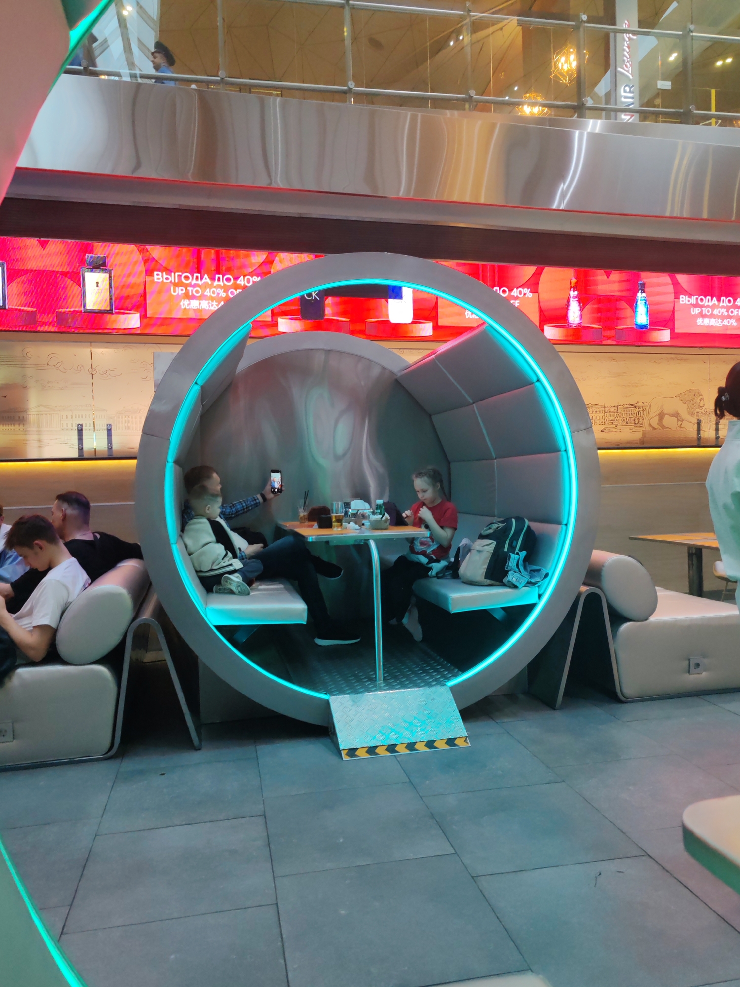 This image depicts a modern, indoor public space, likely a shopping mall or airport lounge in Saint Petersburg, Russia, characterized by contemporary design and functional seating. The central feature is a large, circular, pod-like booth illuminated with bright blue LED strip lighting along its inner circumference, offering a semi-private seating area.
Inside the pod, two adults and two children are relaxing. On the left, an adult man in a plaid shirt is holding a smartphone, seemingly taking a selfie with a young boy seated beside him. They have drinks and a small dish on their table. On the right side of the pod, a young girl in a red shirt is seated at another small table, eating, with a black and grey backpack resting next to her.
Outside the pod, on the left, other individuals are seated in conventional grey upholstered lounge chairs, appearing relaxed. The flooring is composed of large, light grey square tiles.
The background features large digital screens displaying advertisements. Visible text includes "ВЫГОДА ДО 40% / UP TO 40% OFF / 优惠高达40%", indicating a sales promotion, and the brand "CK" (Calvin Klein). Various perfume bottles are shown on the displays. On an upper level, partially visible through a glass balustrade, faint reflections and a person in a cap can be seen. The text "AIR Lounge" is also partially visible on this upper level, suggesting an airport lounge or a travel-themed area within a larger complex. The scene captures a moment of leisure and relaxation within a bustling commercial environment.