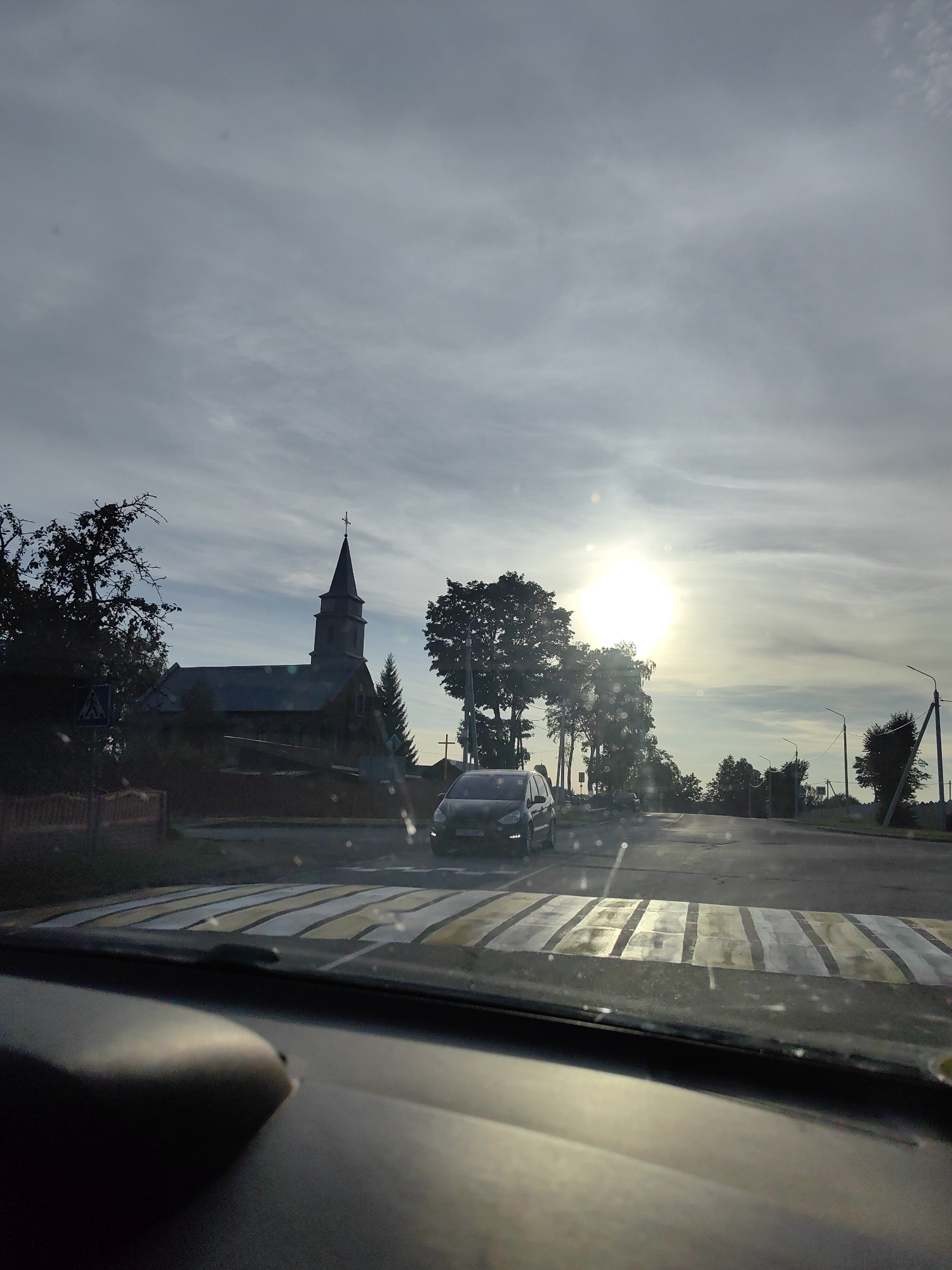 The image depicts a view from inside a vehicle, looking out at a road with a pedestrian crossing. A dark-colored car is stopped at the crossing, waiting. To the left of the road, a dark-colored church with a steeple and a cross is visible, surrounded by trees. The sky is overcast with patches of bright sunlight. The location appears to be a rural or suburban area, consistent with Ilyanskiy Selskiy Soviet, Belarus, based on the provided context. The overall impression is that of a quiet street scene with a church in the background.