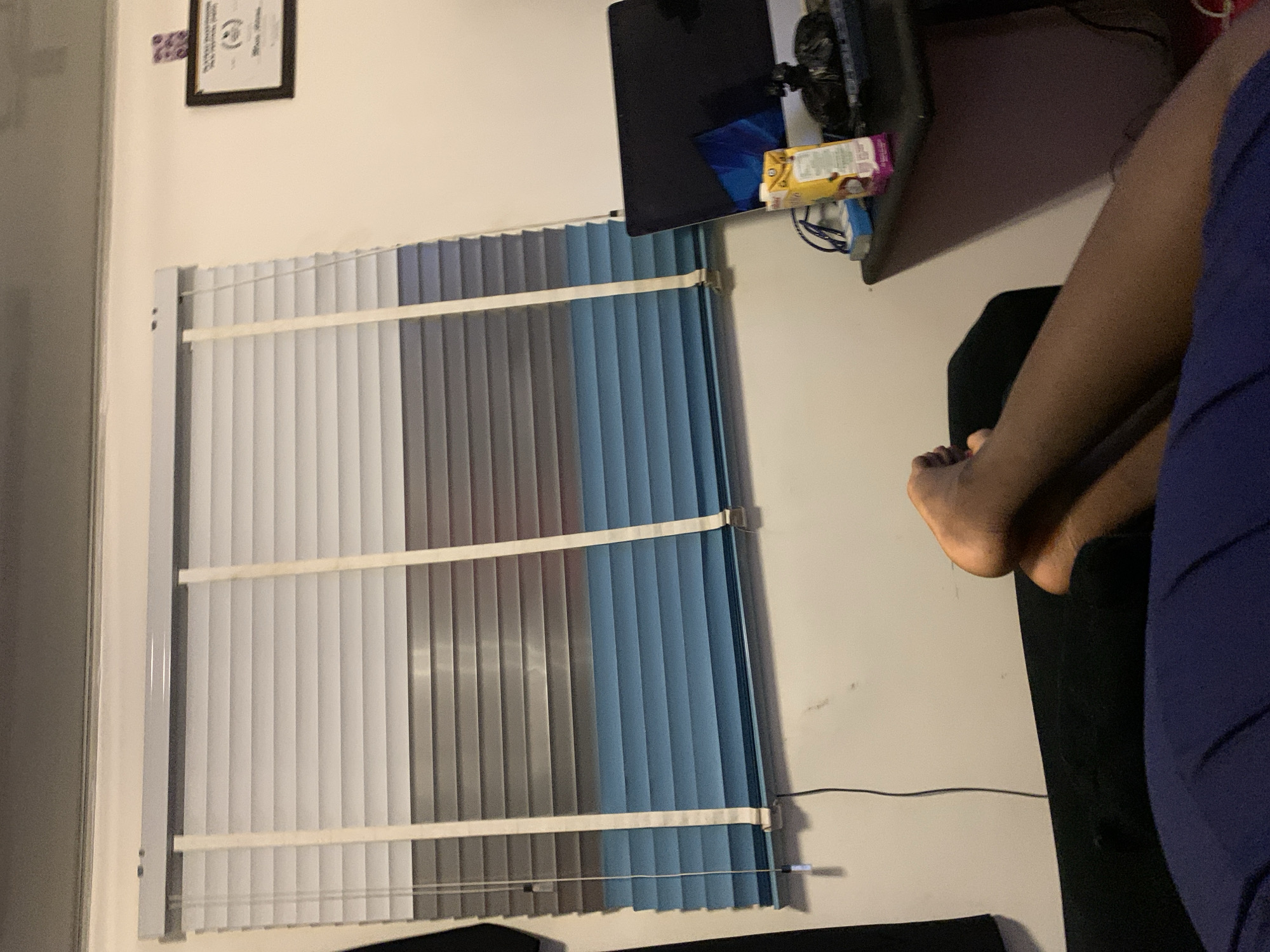 In an indoor scene, a person's bare feet rest on a dark-colored couch, with their legs extending towards the foreground. Their skin tone is dark. The couch is positioned in front of a wall with venetian blinds covering a window. The blinds are a gradient of colors, starting with white at the top, transitioning through shades of gray, and ending in a light blue at the bottom. A black desk is visible to the right of the couch, on which sits a laptop computer and a juice box. The wall above the desk displays a framed document. The overall lighting suggests a dimly lit room.