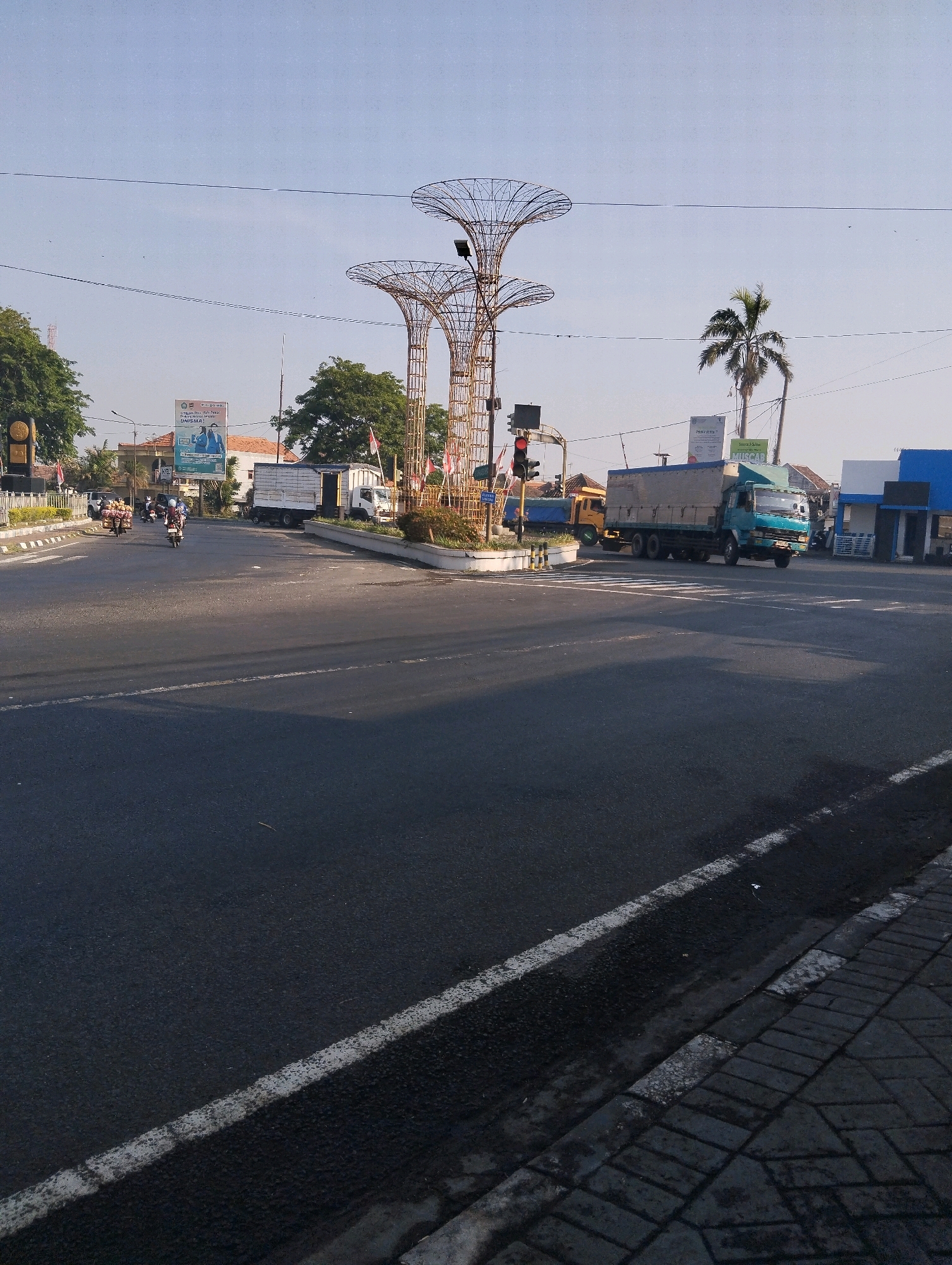 This is a photograph taken at the intersection of Jalan Gatot Subroto and Jalan Dr. Wahidin in Probolinggo, Indonesia. The sun is shining, casting shadows on the road. In the foreground, the asphalt road is visible with white lane markings. To the right, a sidewalk made of interlocking concrete blocks is present. In the middle ground, several vehicles are seen, including a turquoise truck with a silver cargo box, a yellow truck, and several motorcycles. Traffic lights are visible, with one showing red. A distinctive art installation featuring two large, cage-like structures resembling trees stands prominently in the center of the intersection. In the background, buildings, trees, and a billboard are visible. A palm tree is also present on the right side of the frame. The sky is clear and blue.