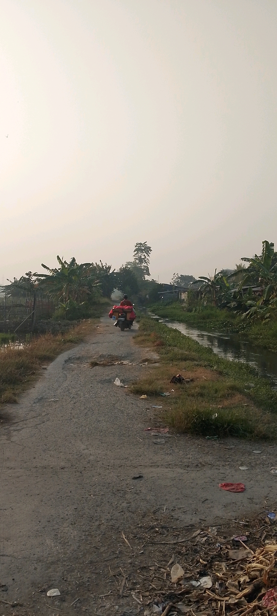 A person is riding a motorcycle loaded with red packages down a dirt road in a rural area, likely Satriajaya, Indonesia. The road is lined with a canal on the right and vegetation, including banana trees, on the left. The sky is overcast, and the overall lighting suggests it is daytime, possibly early morning or late afternoon. Debris, including plastic bags and organic material, is scattered along the side of the road and in the foreground. In the distance, several buildings and trees are visible.