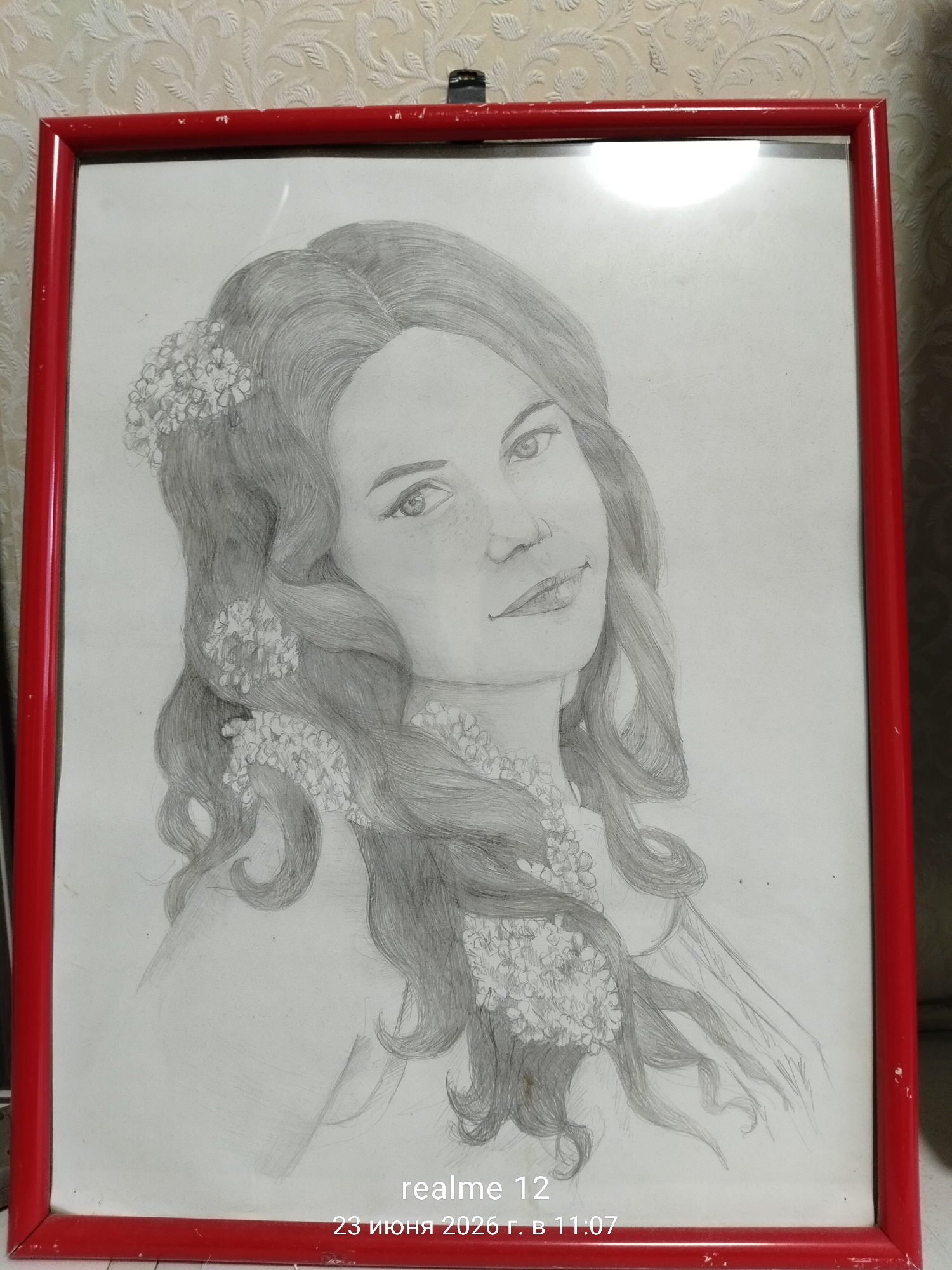 This media file contains a framed pencil sketch of a woman. The woman has long, wavy hair adorned with small flowers. She is depicted from the chest up, with her head tilted slightly and a gentle smile on her face. Her eyes are looking slightly to the side. The sketch is rendered in grayscale, with detailed shading used to create depth and texture, particularly in her hair.
The sketch is enclosed in a red frame. The background of the media file shows a textured wallpaper.
Visible text in the image reads "realme 12" and "23 июня 2026 г. в 11:07". This indicates the device used to capture the image and the date and time of capture, suggesting the photo was taken in Shchyolkovo, Russia. The time of day is noted as 11:07 AM. No specific weather or environmental conditions are depicted. The overall mood of the sketch appears serene and artistic.