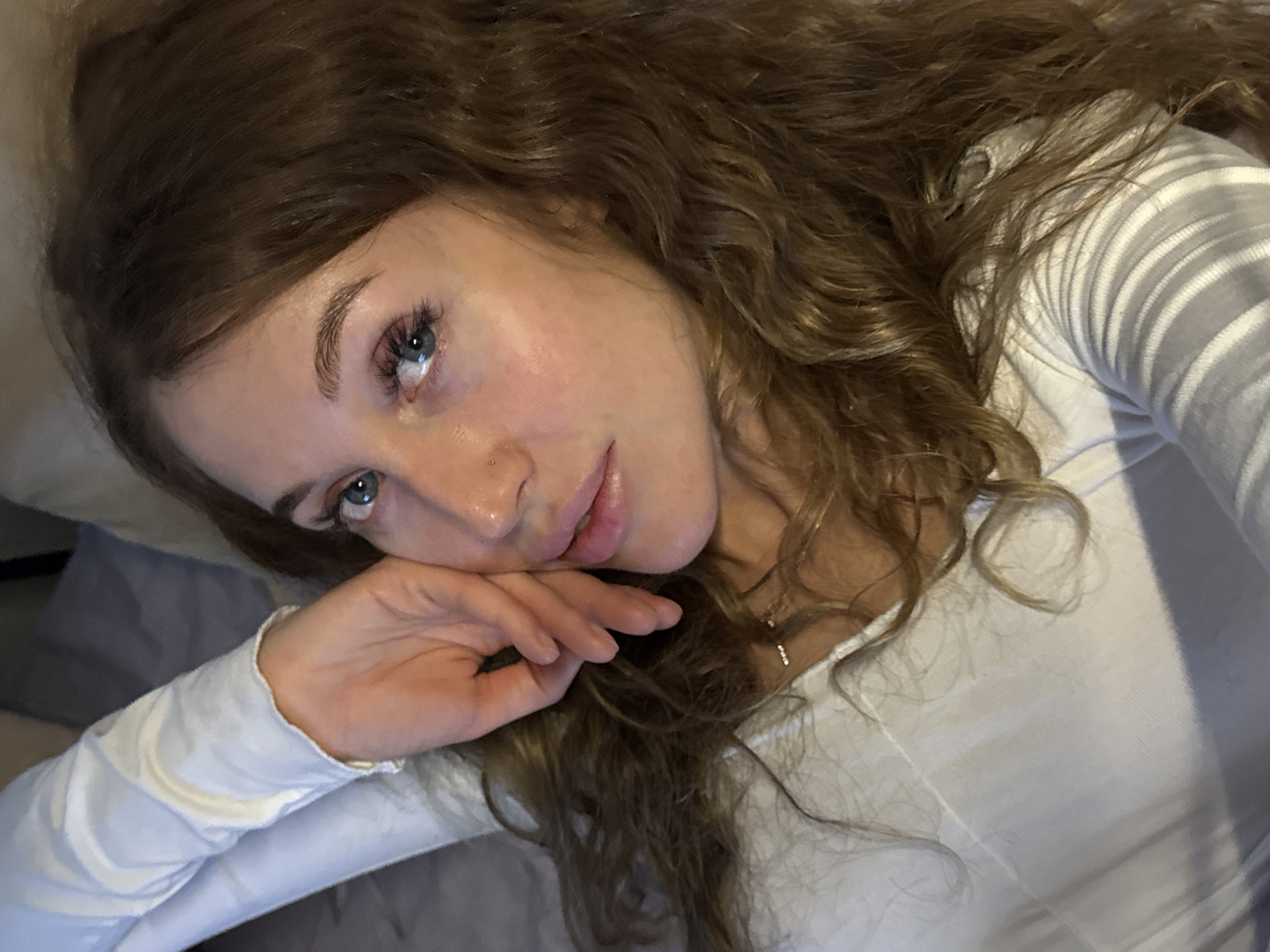 A woman with curly, light brown hair is lying down, resting her head on her hand. She has fair skin, blue eyes, and a small stud piercing in her nose. She is wearing a white, long-sleeved shirt. The background appears to be a bed with light blue sheets. The location is unknown and there is no indication it is in the city of Chaikovsky, Russia.