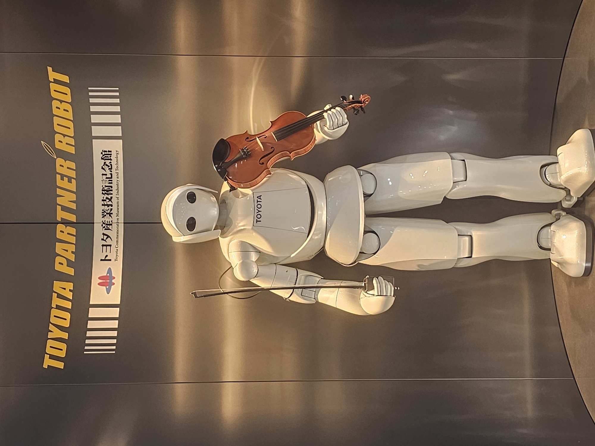 The image depicts a full-body view of a white, humanoid robot standing upright, holding a violin and a bow. The robot has a smooth, featureless head with two dark eye-like sensors. It is dressed in a white, segmented body suit, with visible joints at the shoulders, elbows, waist, knees, and ankles. The violin is positioned across the robot's chest, held by its right hand, while the bow is held vertically in its left hand, near the robot's side.
Above the robot, a large sign with yellow text reads "TOYOTA PARTNER ROBOT". Below this, a smaller sign with Japanese text and the Toyota logo reads "トヨタ産業技術記念館" (Toyota Commemorative Museum of Industry and Technology). The background is a metallic, reflective surface that appears to be part of an indoor exhibition space. The lighting is somewhat warm, casting reflections on the robot and the background.
This robot is located at the Toyota Commemorative Museum of Industry and Technology in Nagoya, Japan. The museum is dedicated to showcasing the history of the Toyota Group's manufacturing and technological innovations.