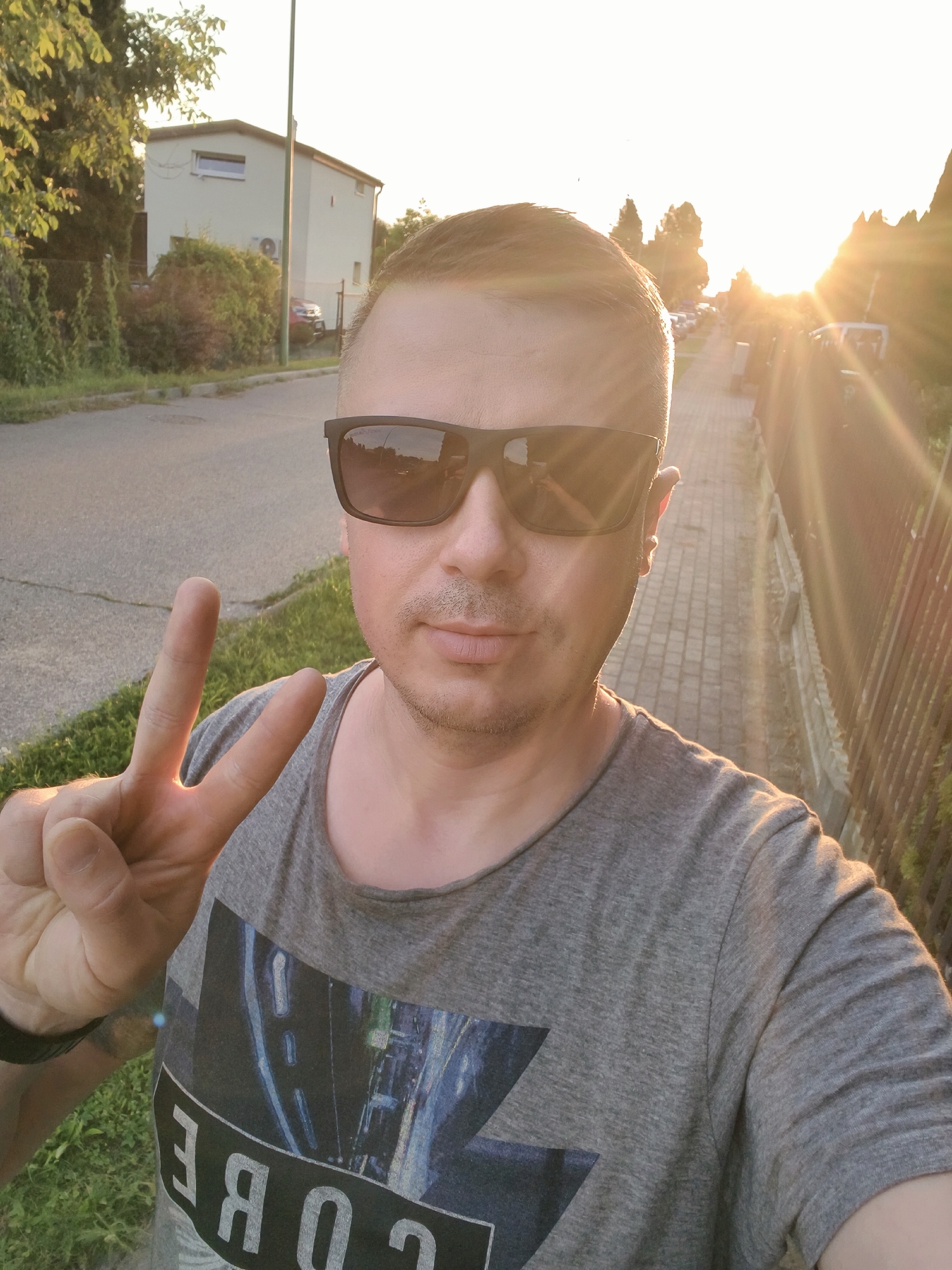 A medium shot captures a man in his 30s wearing sunglasses and a grey t-shirt with a graphic print, holding up two fingers in a peace sign. He is standing on a sidewalk next to a street, with a building and trees visible in the background. The setting sun casts a warm glow and lens flares across the scene. The image appears to be taken in a residential area, likely Tczew, Poland, based on the architectural style and streetscape. The man is the central focus, and his gesture suggests a sense of positivity or a greeting. The overall atmosphere is casual and outdoor.