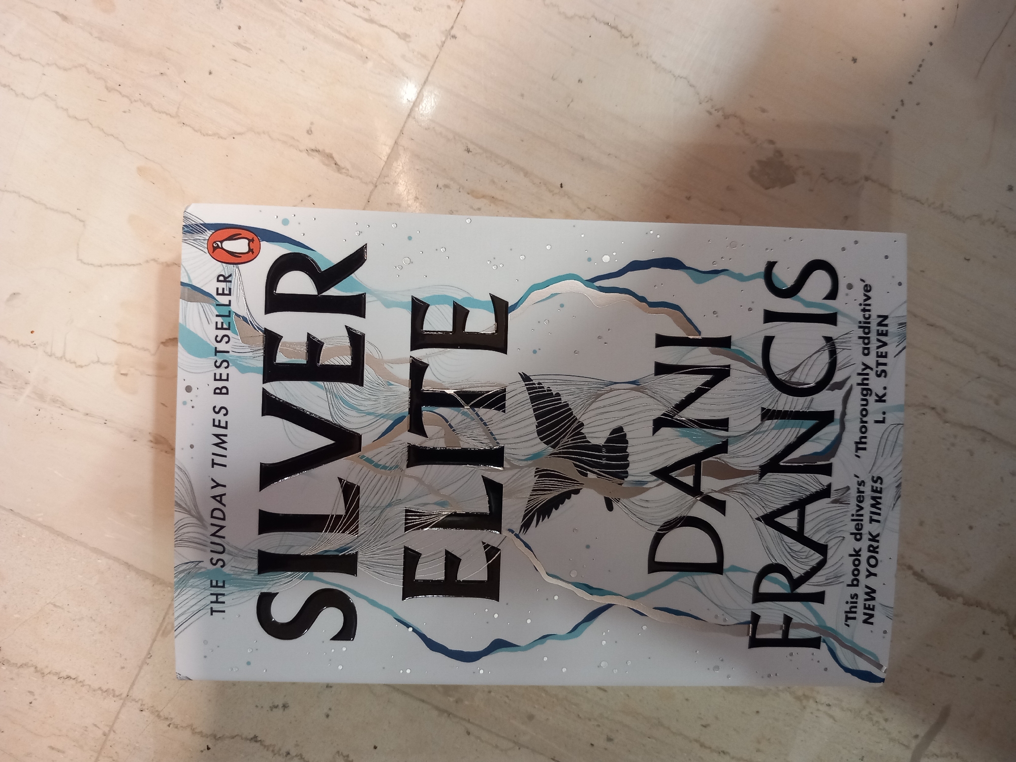 A book with a white cover, featuring the title "SILVER ELITE" in large, black, stylized font, is lying on a tiled surface. The author's name, "DANI FRANCIS," is also visible in black lettering. The cover has a decorative design of abstract blue, beige, and silver lines resembling waves or feathers, with small silver dots scattered across it. A graphic of a bird in black silhouette is positioned between the title and the author's name. In the top left corner of the cover, a red penguin logo with an orange outline, characteristic of Penguin Books, is present. Below the penguin logo, text reads "THE SUNDAY TIMES BESTSELLER." To the right of the author's name, there is a quote from "NEW YORK TIMES" that reads "'Thoroughly addictive' L. K. STEVEN." The book is oriented horizontally, with its spine facing left. The tiled surface appears to be a light-colored, marbled floor.