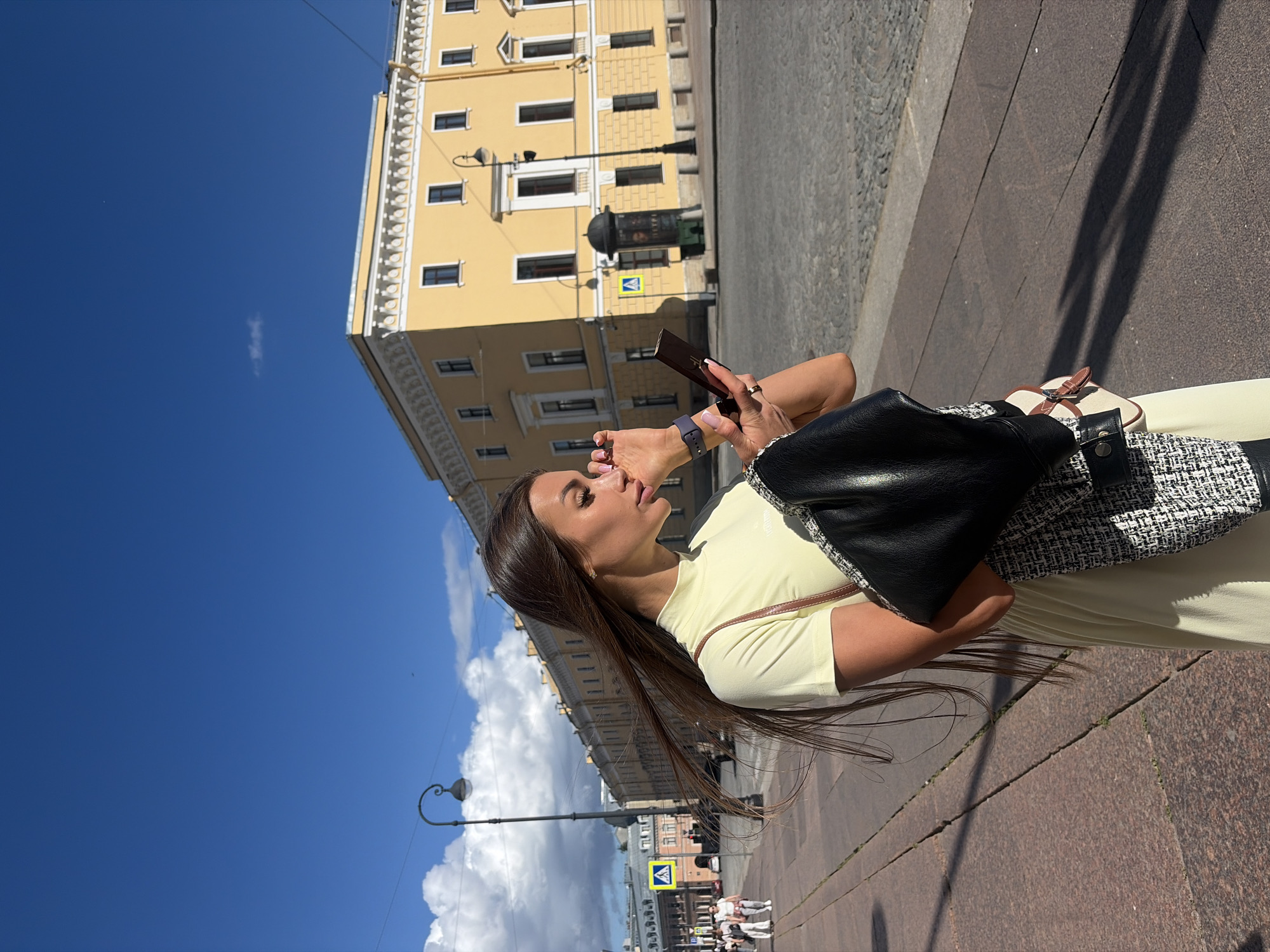 In Saint Petersburg, Russia, under a bright blue sky with scattered clouds, a woman with long, dark hair styled in a high ponytail stands on a cobblestone sidewalk. She is wearing a pale yellow, short-sleeved, high-neck dress and a dark jacket draped over her left shoulder. She holds a compact mirror in her right hand and appears to be applying makeup to her eye, with her left hand touching her face. On her left wrist, she wears a dark smartwatch. A patterned shoulder bag with a black leather flap and brown strap hangs from her right shoulder.
The background features a yellow building with white ornate trim and dark windows. Across the street, a stone-paved area leads to more buildings. A streetlamp with a curved arm stands in the middle ground. Pedestrians are visible in the distance on the sidewalk to the left, and a yellow pedestrian crossing sign is prominently displayed. The scene captures a moment of personal grooming in an urban setting.