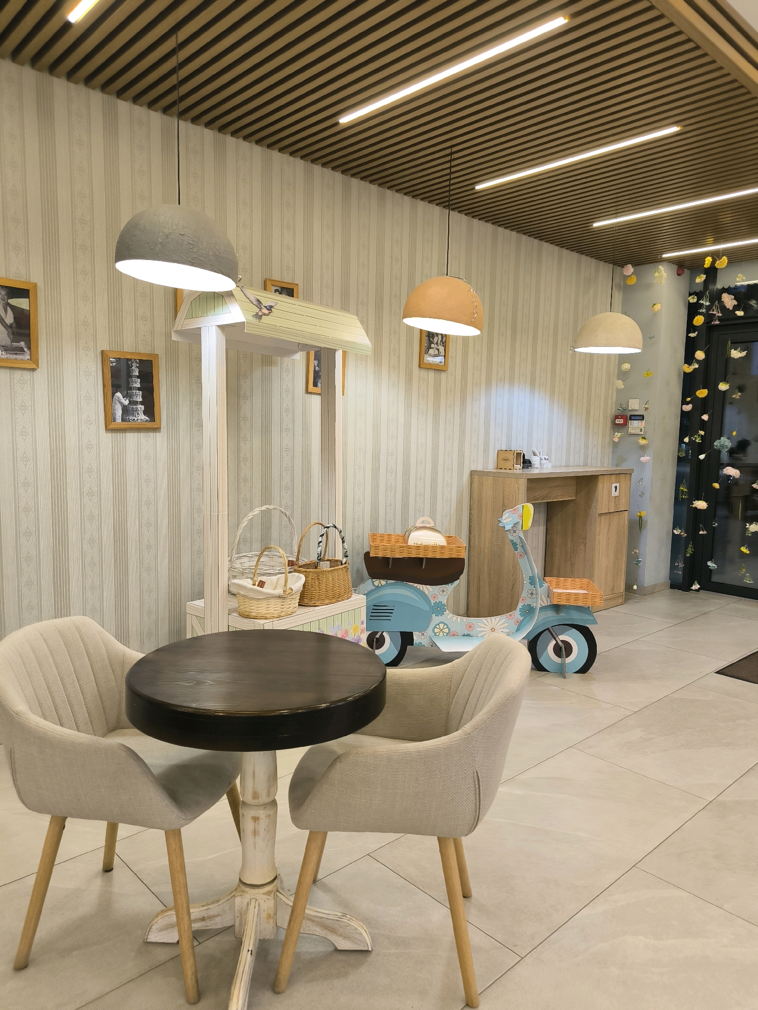 This image shows a quaint cafe interior, possibly located in Chișinău, Moldova. The scene features a circular, dark wood-topped table with a distressed white pedestal base, accompanied by two light-colored upholstered chairs with wooden legs. To the right of the table, a decorative blue toy scooter with floral patterns and wicker baskets is displayed. In the background, a white wooden stand holds several more wicker baskets. The walls are adorned with striped wallpaper and framed pictures. The ceiling is paneled with wooden slats and features modern linear lighting fixtures, along with three prominent pendant lamps. The flooring is tiled with large, light-colored square tiles. The overall ambiance is cheerful and inviting, suggesting a space designed for relaxation and enjoyment.