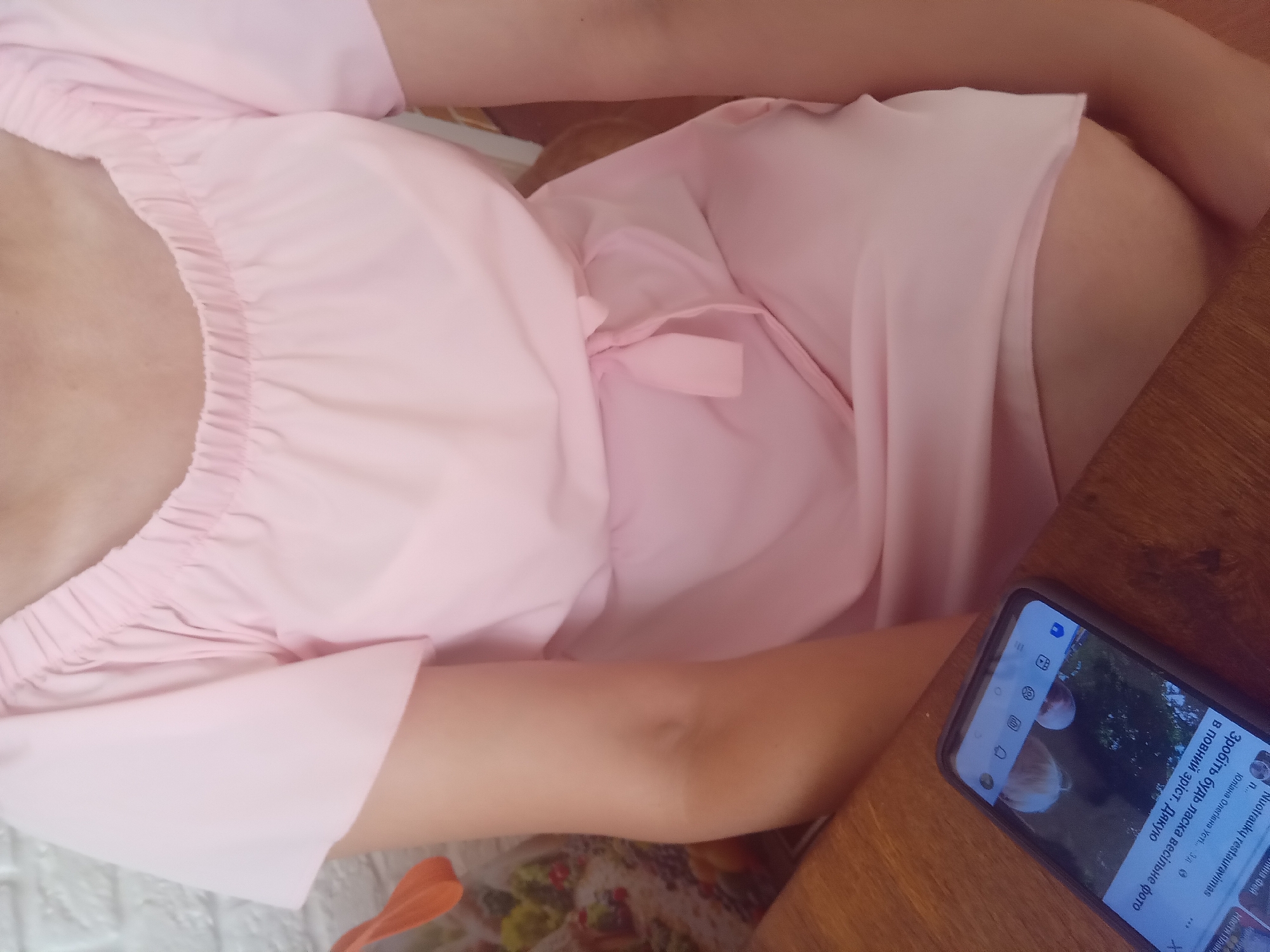 A person wearing a pink dress is seated at a wooden table with a smartphone in front of them. The dress has short, flared sleeves and an elasticized neckline. The smartphone displays social media content, including text in Cyrillic. The background includes a white brick wall and a bag with a floral pattern. The scene appears to be indoors, possibly in a cafe or restaurant in Terebovlia, Ukraine, given the Cyrillic text on the phone.