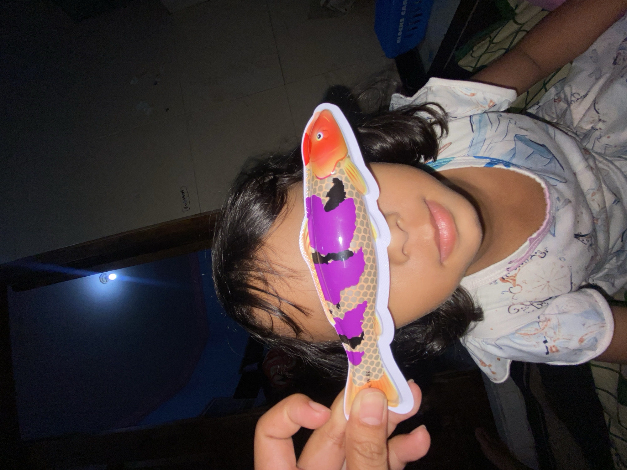 A young girl is lying down, facing sideways, with a colorful, plastic toy fish held up in front of her face. The fish is held by someone out of frame. The lighting is dim, with a light fixture visible on the ceiling. The girl wears a white, patterned top. A blue basket is visible in the background. The setting is indoors. The location is potentially in the city of Gelam, Indonesia.