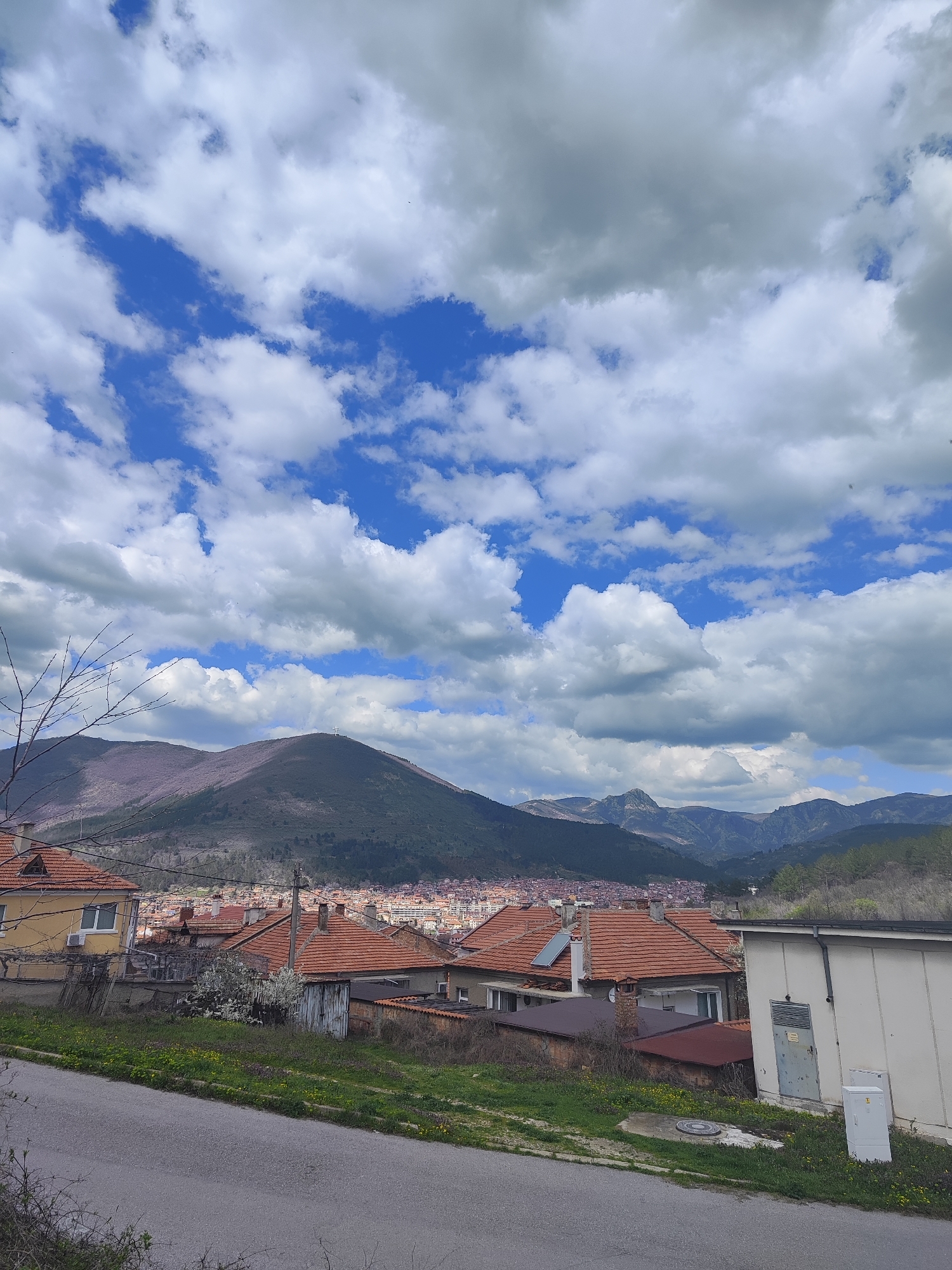 The image captures a view in Sliven, Bulgaria, showcasing a residential area against a backdrop of mountains and a partly cloudy sky. The scene is composed of a street in the foreground, transitioning into a neighborhood with houses, most of which have orange-tiled roofs. Behind the houses, a mountain range stretches across the horizon, partially obscured by atmospheric haze. The sky is dominated by a mix of white and gray clouds, interspersed with patches of blue sky.
Key features include:
* **Buildings:** Houses with red tile roofs are prominent in the mid-ground, indicating a residential area. A white building stands on the right.
* **Environment:** The foreground features a paved road and greenery, with yellow flowers scattered among the grass.
* **Mountains:** In the background, a mountain range stretches across the horizon.
* **Sky:** The sky shows a mix of white and gray clouds, with patches of blue.
* **Vegetation:** Trees and other plants can be seen on the hillside and in gardens.