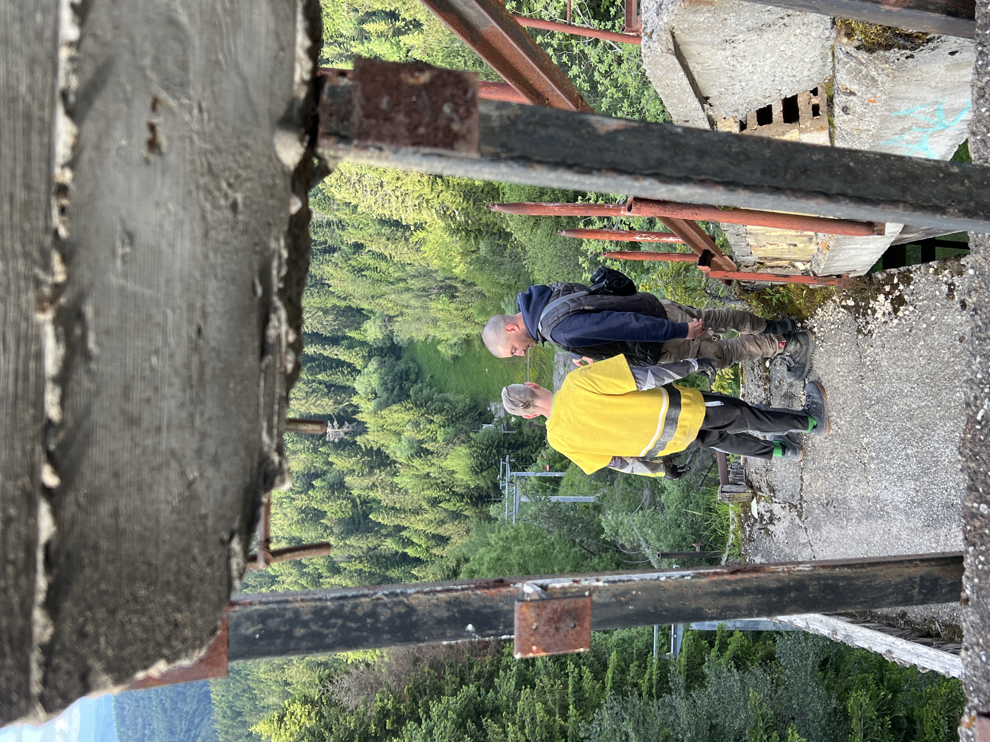 Two men are standing on the edge of a decaying concrete structure, overlooking a densely forested valley. The man on the left wears a bright yellow t-shirt with reflective stripes, black pants, and green socks. The man on the right is dressed in a dark vest over a blue hoodie, khaki pants, and dark boots, with a backpack slung over his shoulder. The concrete structure appears to be an abandoned elevated platform or bridge, with rusted metal railings and broken concrete edges. Below them, a ski lift or similar aerial transportation system is visible amidst the trees, and the landscape extends into rolling hills under a bright sky. The overall scene suggests an exploration of an abandoned industrial or recreational site in a natural environment.