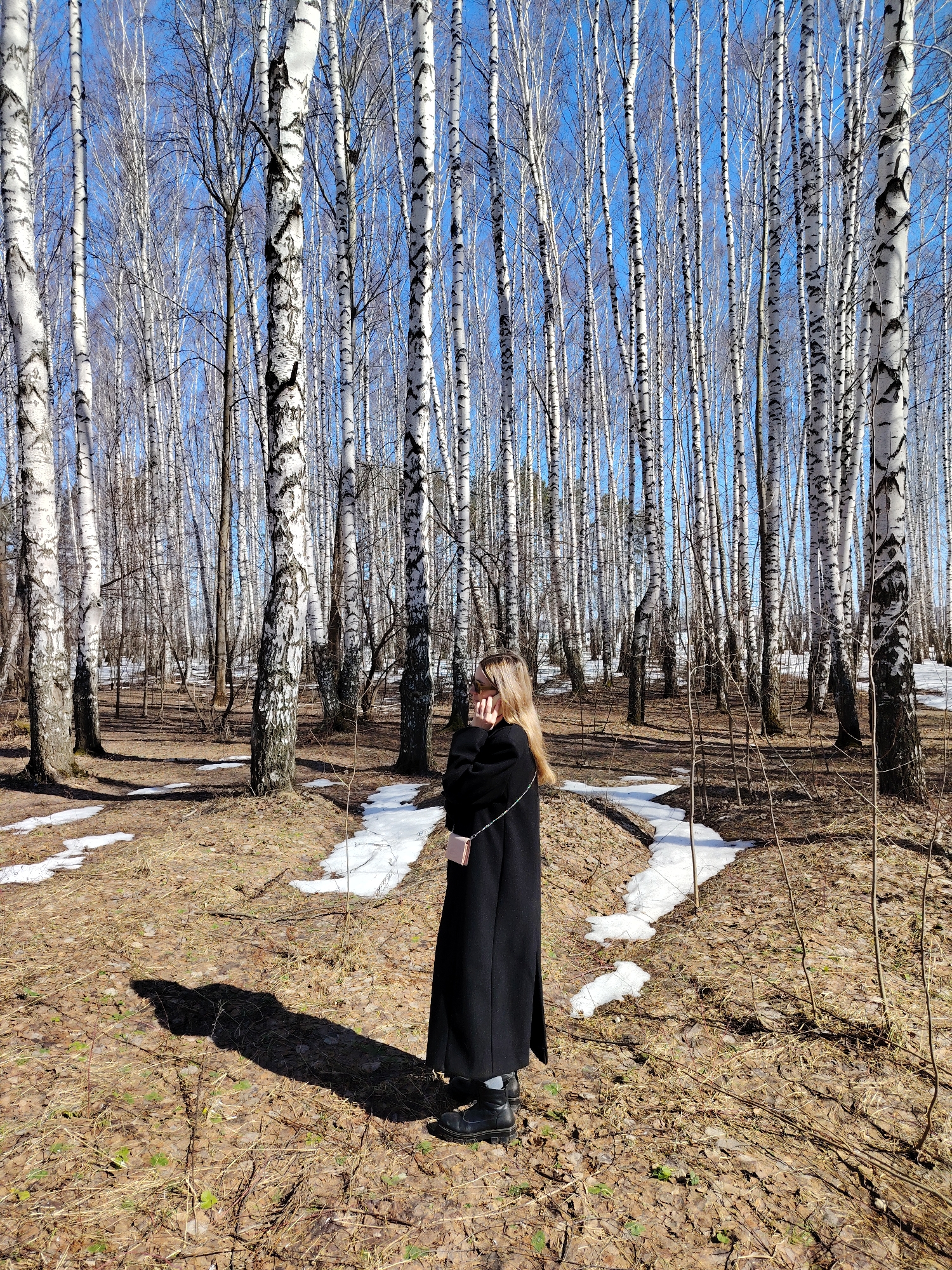 The image depicts a woman standing in a birch forest. The woman is wearing a long black coat and dark shoes. She is holding a mobile phone to her ear with her right hand. The forest floor is covered with brown leaves and patches of melting snow. The sky is blue and the trees are bare, indicating that it is early spring. Based on geotagging, the location is likely in or near the city of Chaikovsky, Perm Krai, Russia, given the presence of a birch forest characteristic of that region.